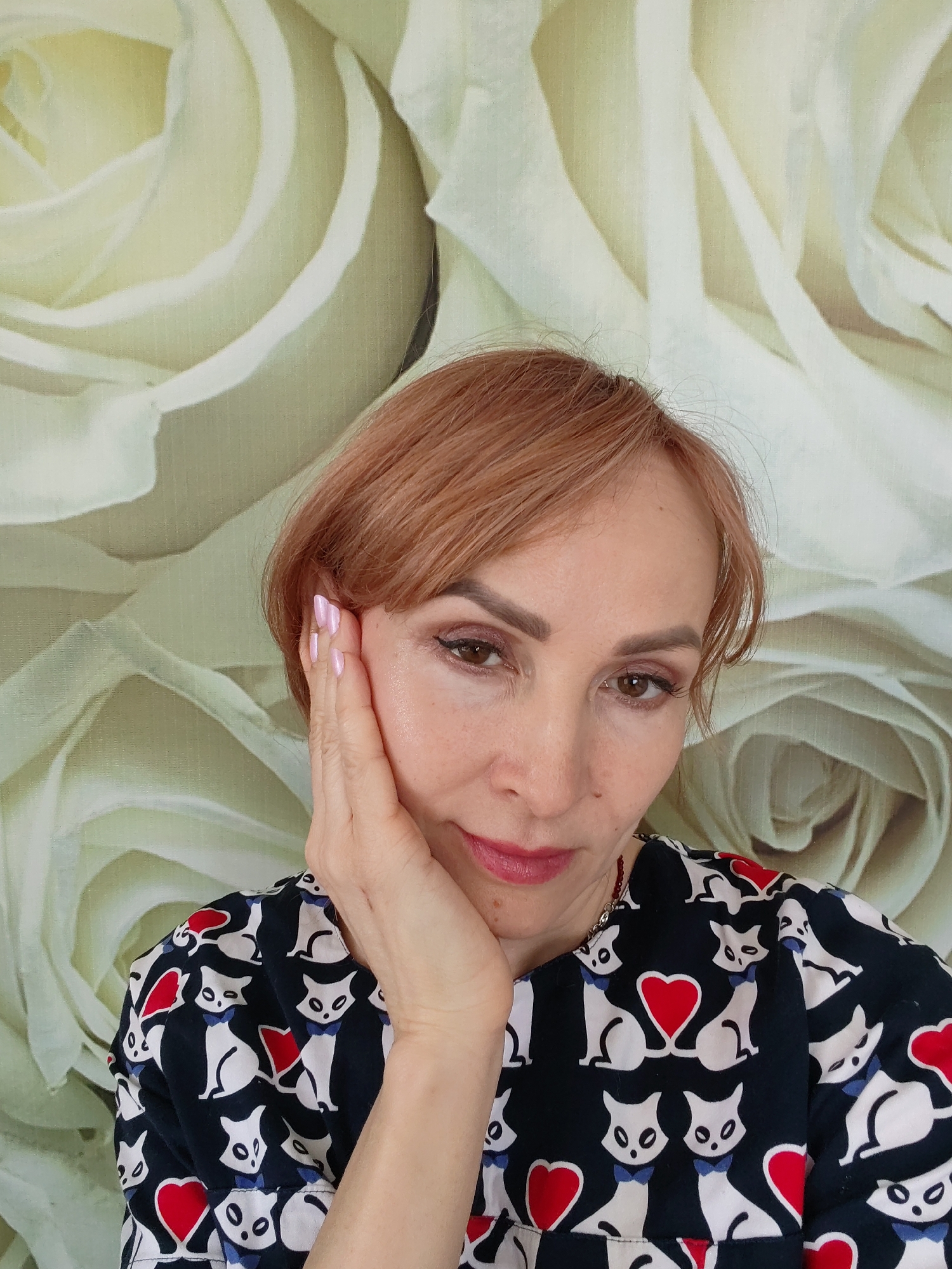 A close-up portrait shows a middle-aged woman, likely indoors in Chaikovsky, Russia. She has light brown or auburn short hair with bangs, dark brown eyes, and is wearing visible makeup including eyeliner, eyeshadow, and light red lipstick. Her right hand, adorned with pink nail polish, gently rests against her cheek and jawline. She is wearing a dark-colored top featuring a playful pattern of white cats with blue bow ties and red hearts. The background is dominated by a large, soft-focus image or wallpaper displaying delicate white and cream roses. The woman maintains a calm and direct gaze towards the viewer. No text is visible in the image.