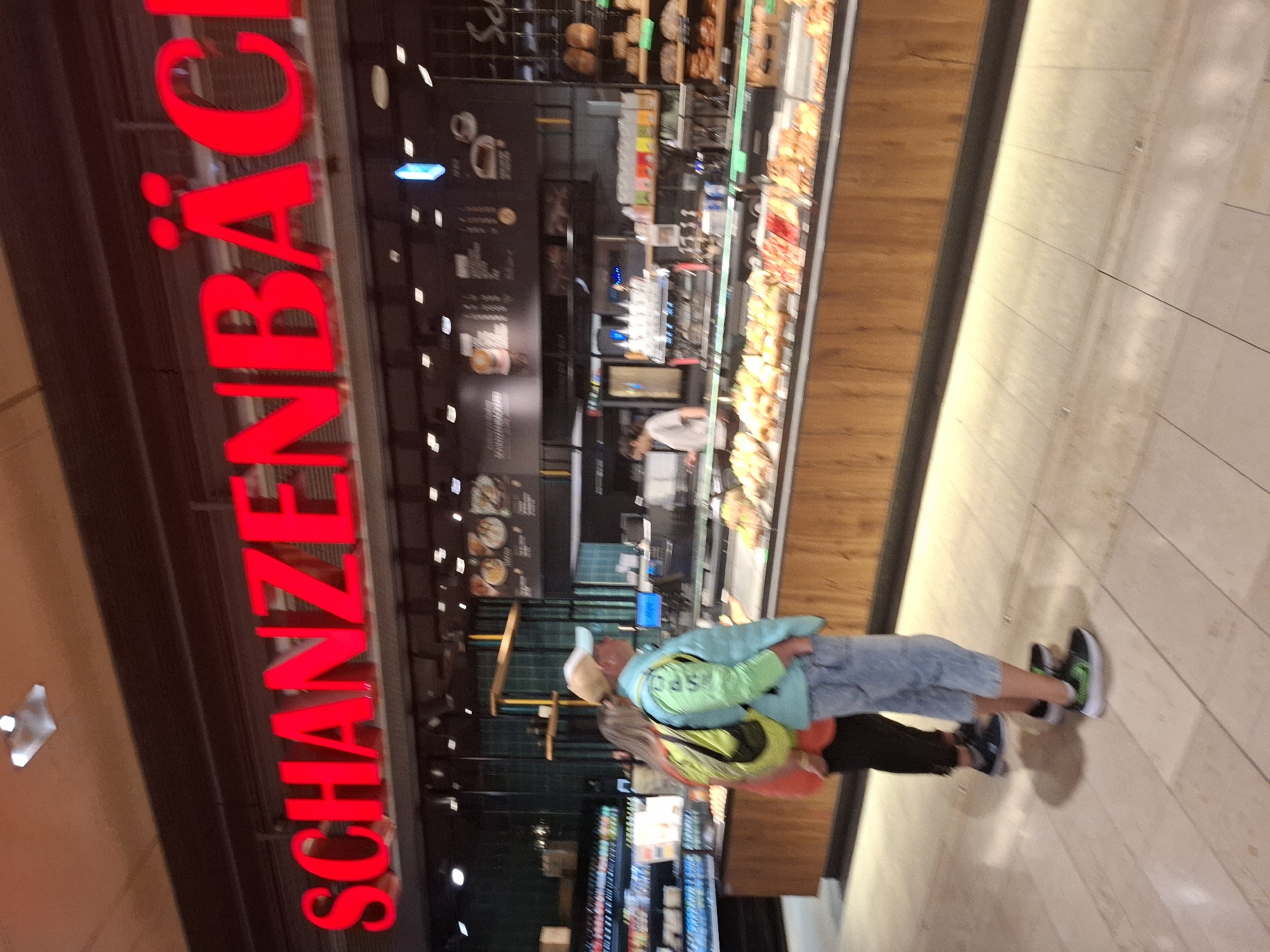 The image captures a storefront with a large, illuminated red sign that reads "SCHANZENBÄCKEREI." This sign is prominently displayed at the top of the image. Below the sign, a bakery display counter is visible, filled with various pastries and baked goods. In the foreground, two individuals are standing and facing the counter, appearing to be customers. One person, seen from the back, is wearing a bright orange top and a yellow backpack. The other person, facing to the right, is wearing a light blue vest over a light green long-sleeved shirt, a baseball cap, and light-colored trousers. The environment appears to be a well-lit interior, likely a bakery or a food establishment within a shopping area or train station. The signage and name suggest the location is within Hamburg, Germany, specifically the Schanzenviertel, a well-known district in the city.