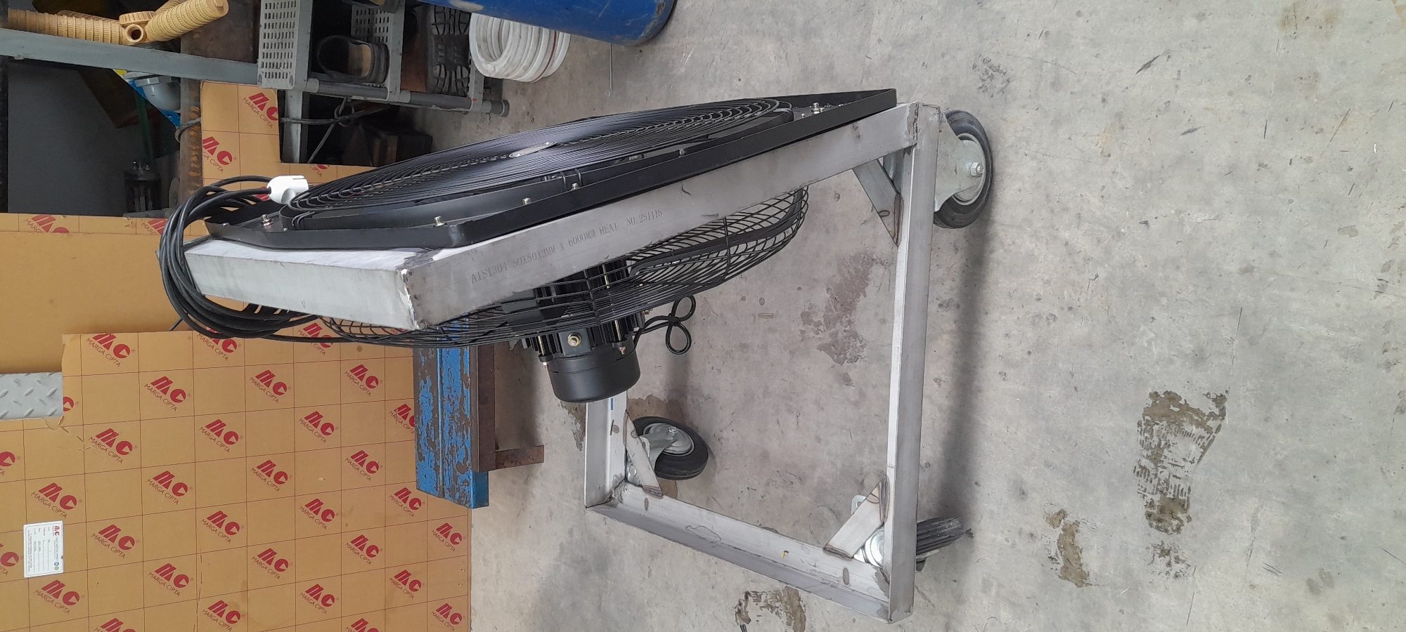 A black industrial fan, mounted on a metal wheeled cart, is the primary subject of the image. The fan's cage is visible, and the motor is a dark cylindrical shape. The cart has a simple rectangular frame with four wheels, two of which are clearly visible and appear to be castors. The fan is tilted upwards at an angle. In the background, cardboard boxes with red lettering that reads "Marga Cipta" are stacked. A blue wooden bench is also visible behind the cart. The floor is concrete, with some scuff marks and what appear to be shoe prints. The overall environment suggests an indoor industrial or workshop setting.