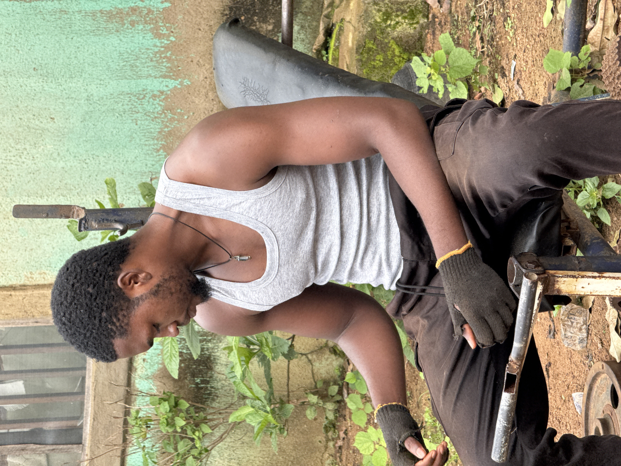 A young Black man with a beard is sitting on a weight bench outdoors, facing to his left. He is wearing a grey tank top, dark brown sweatpants, and black fingerless gloves with yellow trim. He has a necklace with a clear pendant. His left arm is resting on his thigh, and his hands are loosely clasped. The bench appears to be made of black material and is supported by a rusty metal frame. In the foreground, there is a weight plate on the ground. Behind the man, there is a light green painted wall with a window visible to the left. The ground is covered in dirt and scattered leaves, with green plants growing around the bench. The overall setting suggests an outdoor gym or a casual workout space. The image was taken in Akwanga, Nigeria.