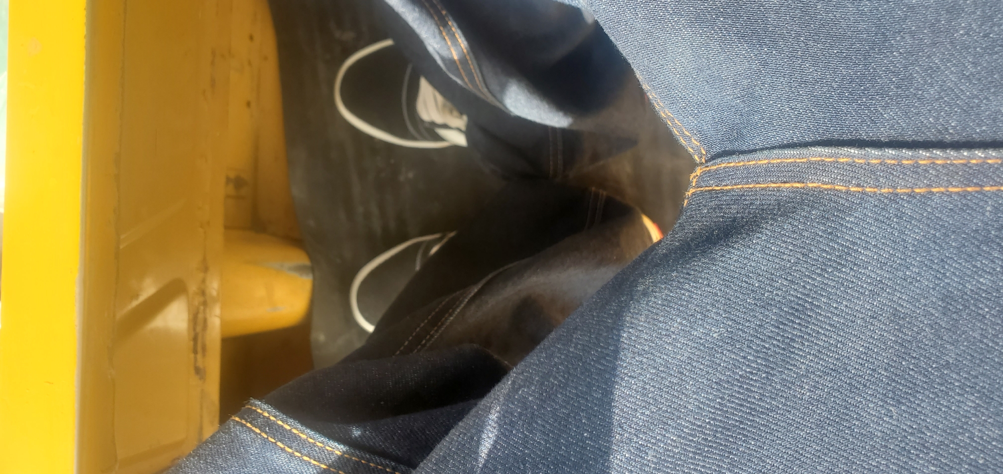 The image shows a close-up perspective of a person's legs wearing blue denim jeans and black and white sneakers. The person is seated, as indicated by the angle of the legs and the presence of what appears to be a seat or platform beneath them. The background features a bright yellow surface, possibly part of a structure or seating, with some weathered areas and a yellow cylindrical object. The scene is lit by sunlight, casting shadows and highlighting the texture of the denim. The overall impression is casual and informal. There is no discernible information in the image to indicate its location in the city of Jos, Nigeria.
