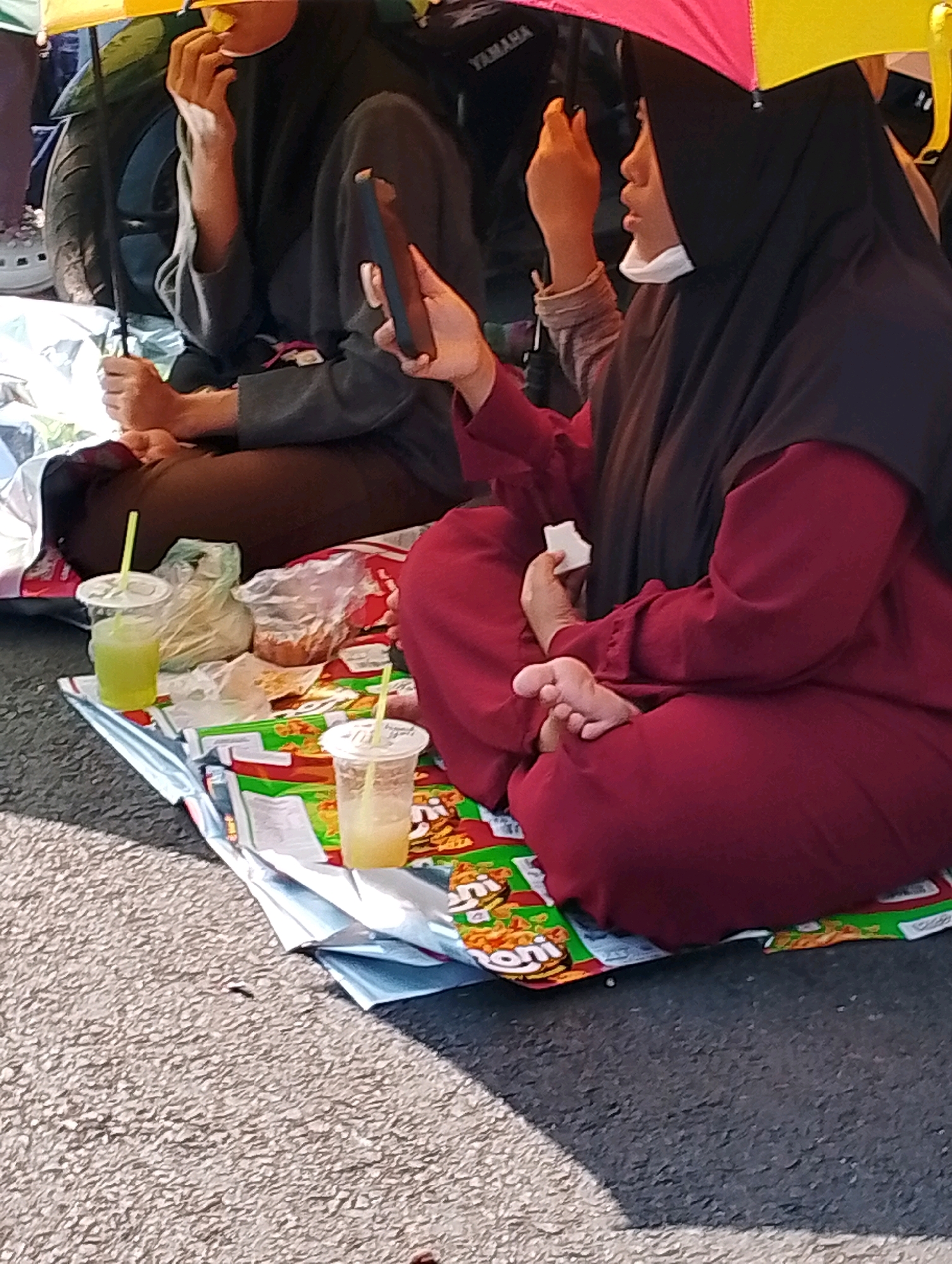 The image shows a group of people sitting on a blanket or mat on a paved surface, likely a street, in Probolinggo, Indonesia. The primary focus is on two individuals. One person on the right is wearing a dark hijab and a maroon abaya, holding a smartphone in their right hand, and is partially obscured by a colorful umbrella. The other person, in the center, is wearing a dark gray long-sleeved shirt and brown pants, and is holding a piece of food near their mouth. Both individuals appear to be looking down or at their phones.
In the foreground, there are two plastic cups with straws, filled with a light yellow liquid, possibly juice. Adjacent to the cups are some crumpled plastic bags and what appears to be packaged snacks. The background shows part of a motorcycle and a hint of other people. The overall impression is of a casual gathering or rest stop on a public road, with individuals engaged with their personal devices and consuming refreshments. The bright, likely sunny, weather is suggested by the umbrella and the overall lighting.