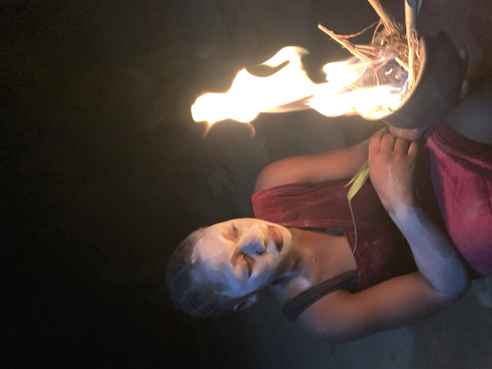 A person with white paste on their face lies prone in a dark environment, partially illuminated by a flame emanating from a container held by another individual. The subject wears a red garment. Location cannot be identified.