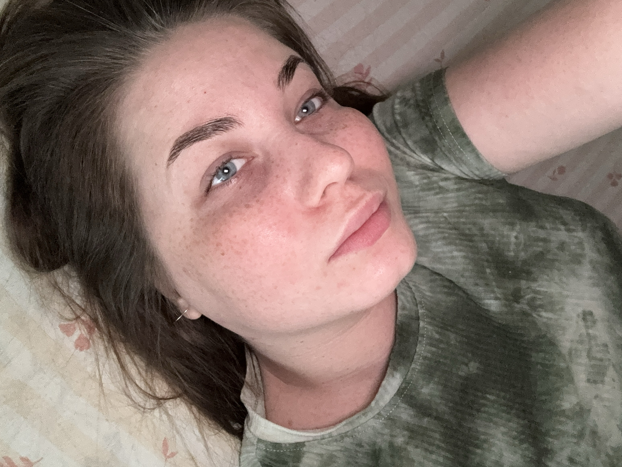 A woman with brown hair and fair skin with freckles, wearing a green and gray tie-dye shirt, is lying on a light-colored surface with a floral pattern. Her head is angled towards the camera, and she is looking up with light blue eyes. She is wearing a small hoop earring in her left ear. The location is not identifiable as Chaikovsky, Russia.