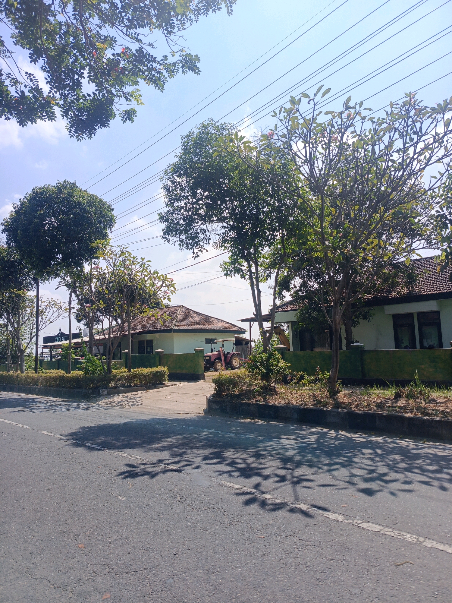 The image shows a residential scene on a sunny day in Tambran, Indonesia. In the foreground, a paved road with a white line marking the edge is visible, with shadows of trees cast upon it. Beyond the road, there is a well-maintained lawn and a low, green-painted fence. Behind the fence, two single-story houses with tiled roofs are situated. The house on the left has a more visible entrance area with a roof supported by poles, and a sign that reads "CBD 24". The house on the right has a similar architectural style. A red tractor is parked in the driveway of the house on the right. Several trees are scattered throughout the property, with lush green foliage. Overhead, power lines stretch across the clear blue sky, with some scattered white clouds. The overall impression is one of a peaceful, suburban or rural residential area.