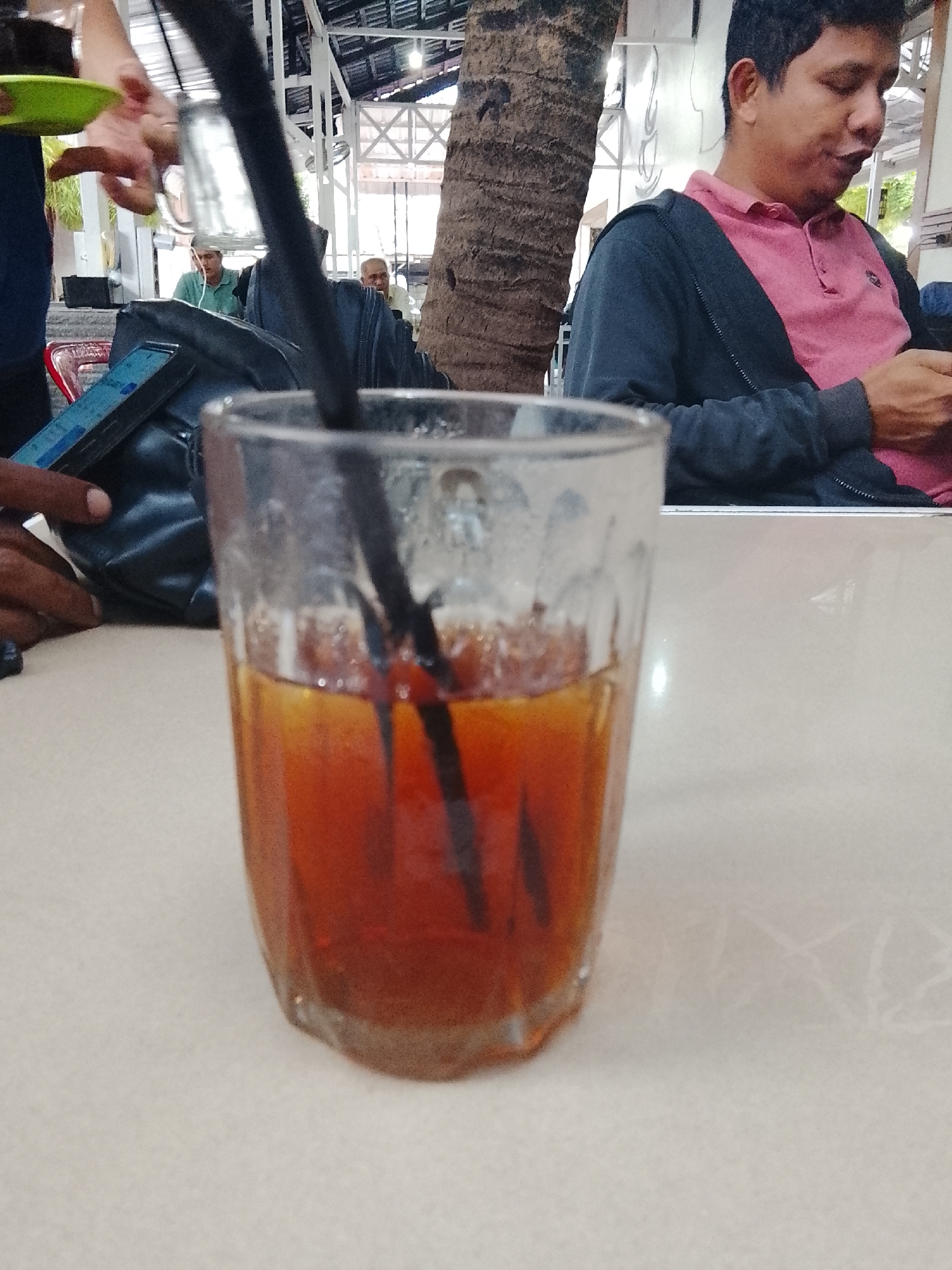 The image shows a glass of iced tea with two black straws on a light-colored table, with several people in the background. One person, wearing a pink shirt and dark jacket, is looking at a phone. A black bag and a hand holding a smartphone are visible to the left of the glass. The background includes other people, a palm tree trunk, and elements of what appears to be an outdoor dining area with a roof structure. The location is likely in Lhokseumawe, Indonesia.