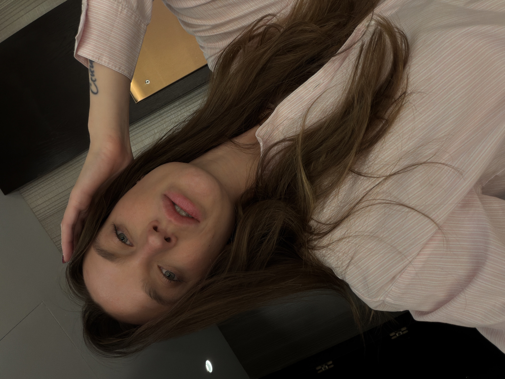 A woman is lying on her side. She has long brown hair and is wearing a pink and white striped shirt. Her hand is touching her forehead. A tattoo is visible on her inner arm. A black surface and a gold rectangular object appear in the upper left corner. A light is visible on the floor in the lower left corner. The image provides no information about the city of Budva, Montenegro.