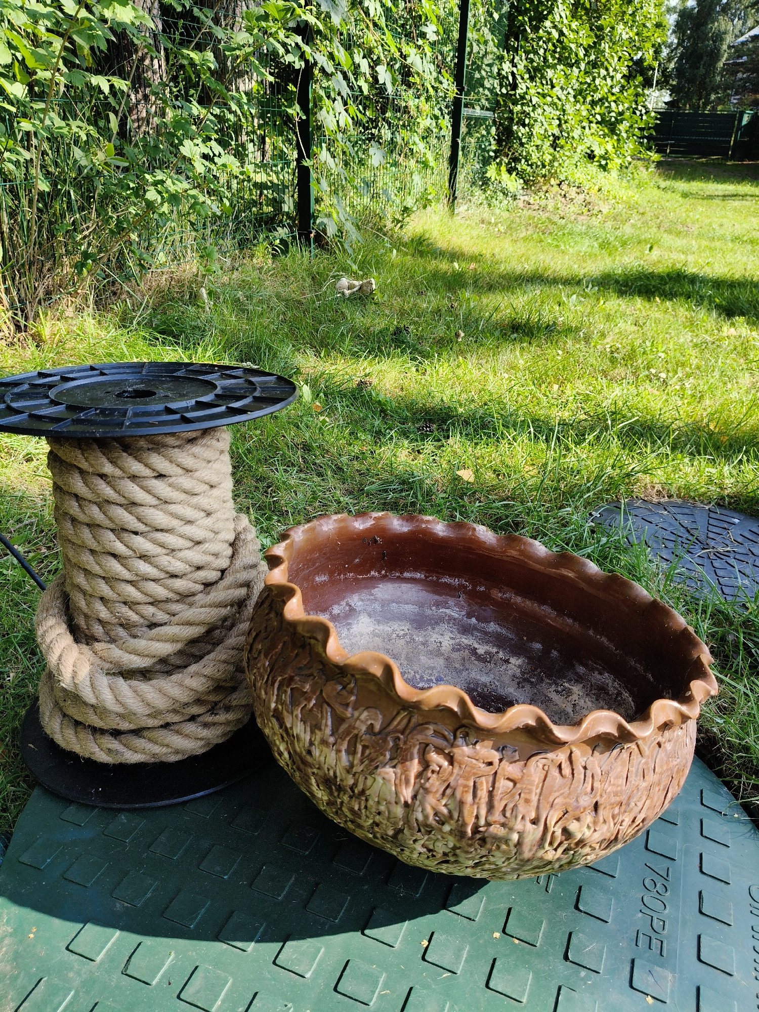 In Bigauņciems, Latvia, a spool of thick, beige rope is positioned on a green, patterned mat, with a decorative brown ceramic planter to its right. The planter has a ruffled rim and an intricately textured exterior. Behind the objects, a well-maintained grassy area transitions into a dense green hedge bordering a metal fence. Sunlight casts shadows on the grass, and the overall scene suggests an outdoor setting, possibly a garden or backyard.