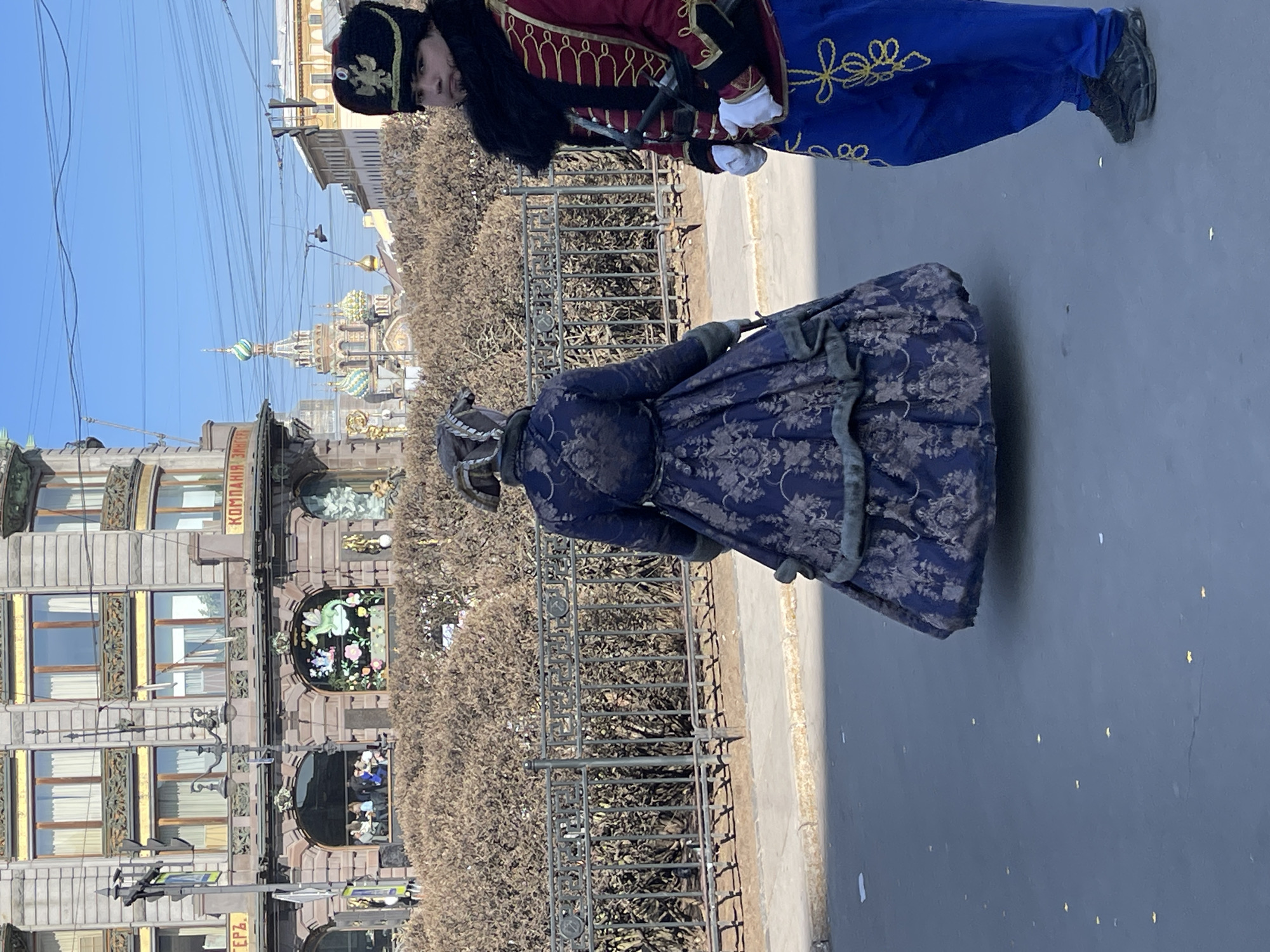 The image depicts a street scene in Saint Petersburg, Russia, featuring two individuals dressed in period costumes. The man is wearing a red and blue military-style uniform with gold embellishments and a fur hat. The woman is in a dark blue floral patterned dress with silver trim. They are standing on a sidewalk next to a metal fence with bushes behind it. In the background, the Singer House and the Church of the Savior on Spilled Blood are visible. The sky is clear and blue.