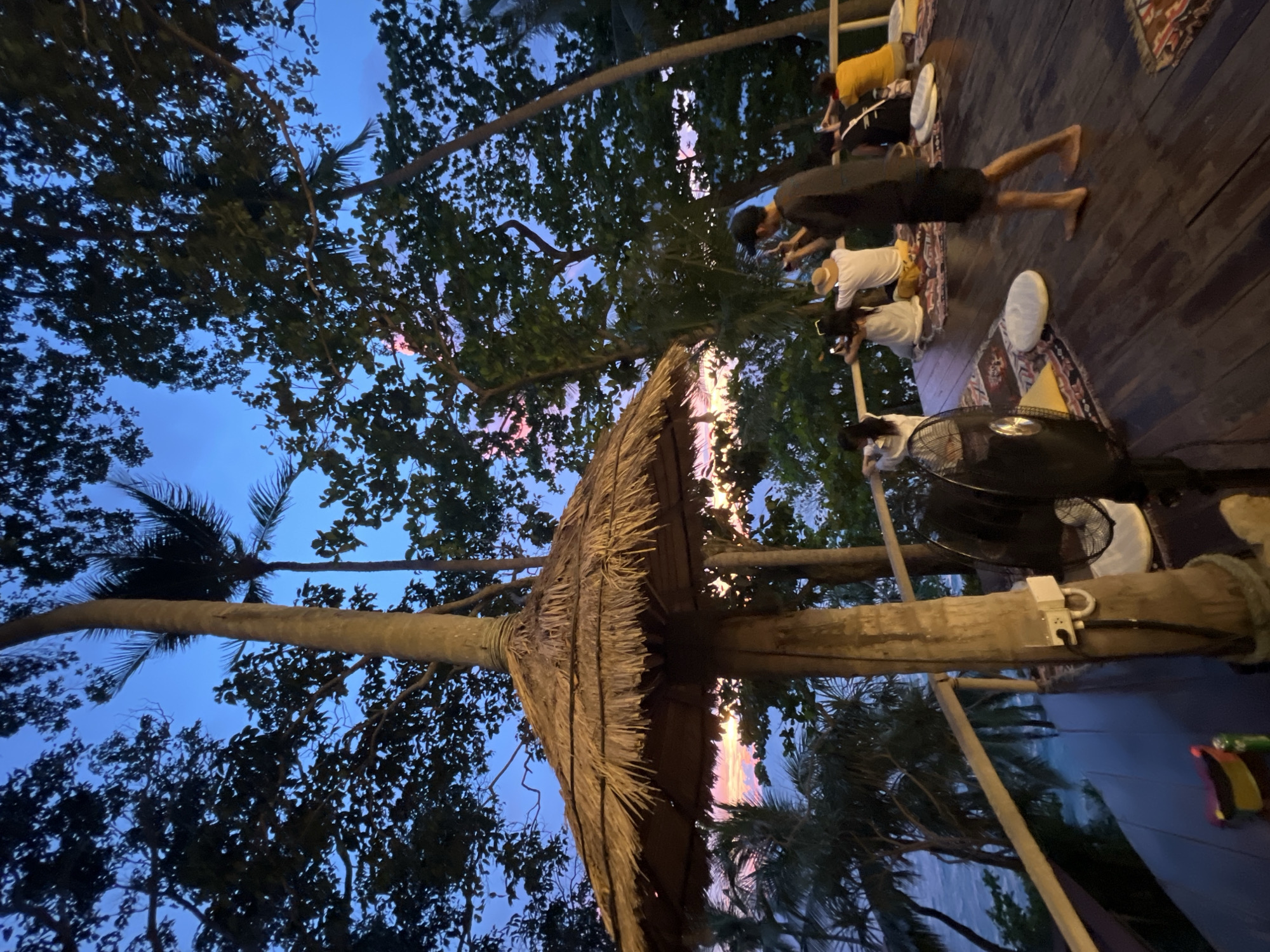 The image depicts a group of people sitting on a wooden deck overlooking the ocean at dusk in Karon, Thailand. A large, thatched umbrella stands prominently in the foreground. Several individuals are seated on the deck, facing the water and holding up their phones, suggesting they are taking pictures or videos of the sunset. Palm trees and dense foliage frame the scene, with the sky displaying hues of blue, pink, and orange. A fan is visible on the left side of the deck, and colorful rugs and cushions are scattered around for seating. The overall atmosphere appears relaxed and scenic, capturing a moment of enjoyment by the coast.