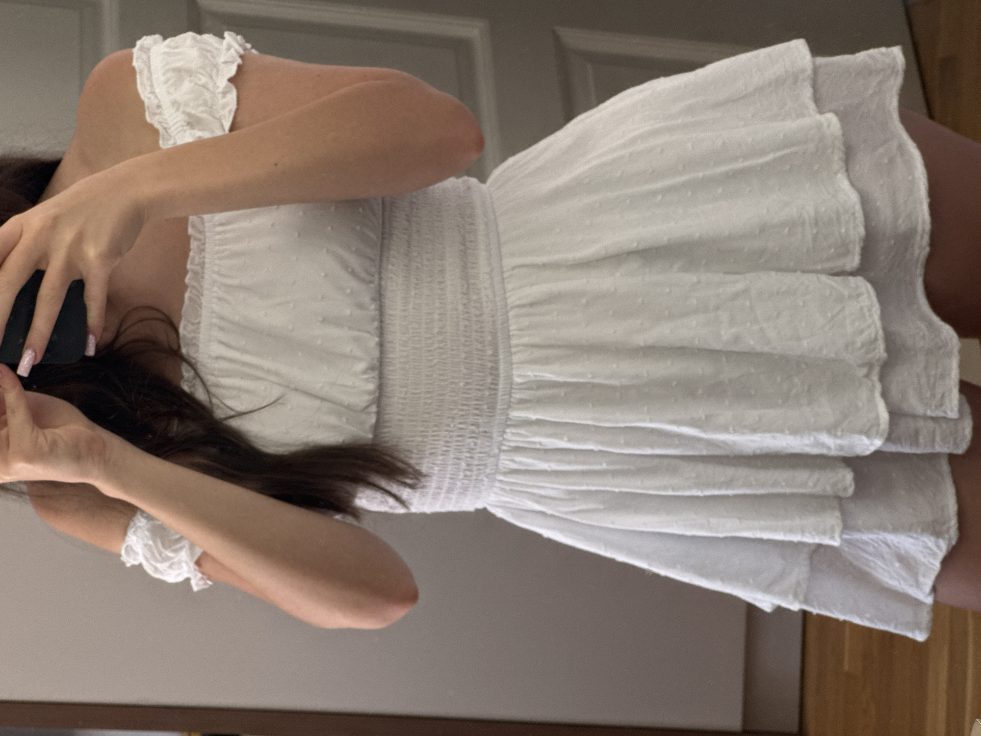 A person stands in front of a mirror, taking a picture of themselves. They are wearing a white, off-the-shoulder dress with smocking around the waist. The dress has a tiered skirt and puffed sleeves with ruffled edges. The background shows a wooden floor and a closet door.