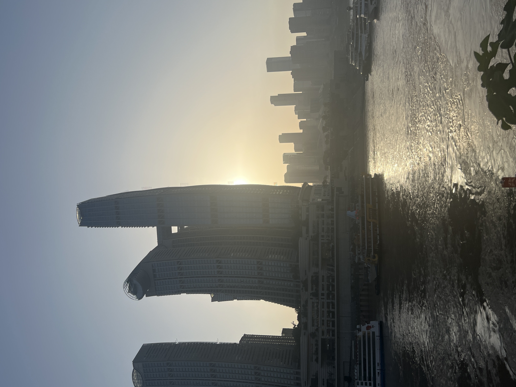The image depicts a city skyline at sunset or sunrise, viewed from a body of water. Prominent in the foreground are several modern skyscrapers, with one particularly distinctive building featuring a curved, angled structure topped with a circular element and another section that appears to be a bridge connecting two towers. The buildings are silhouetted against a hazy sky with the sun low on the horizon, casting a golden glow. The water in the foreground reflects the light and the cityscape, with ripples indicating movement. Several boats, including what appear to be passenger ferries, are visible on the water, some docked and others in motion. In the mid-ground and background, a denser collection of buildings forms the rest of the city's silhouette, partially obscured by atmospheric haze. The scene suggests a bustling urban environment, likely a port city, with the Nan'an District in Chongqing, China, being a likely location given the characteristic architecture of the Yangtze River. There are no discernible people in the image.