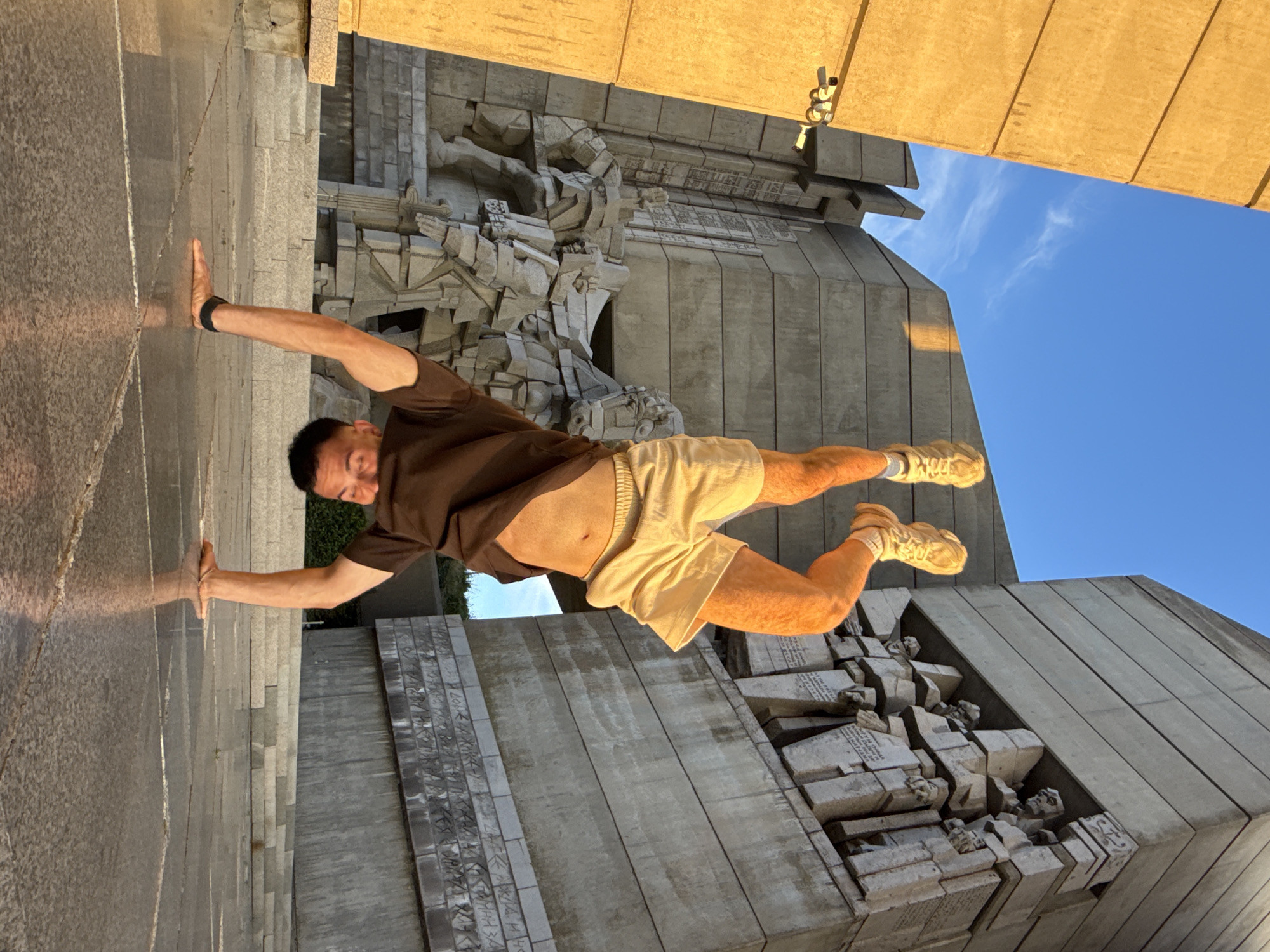 A man performs a handstand on a paved plaza in front of the Monument to the Declared Founders of the Bulgarian State in Shumen, Bulgaria. The monument, constructed from concrete, features relief sculptures depicting historical figures. The man, wearing a brown t-shirt, light-colored shorts, and yellow sneakers, is upside down with his legs extended upwards. The sun casts a warm glow on the scene, highlighting the textured surfaces of the monument and the wet pavement. A security camera is visible on a nearby concrete structure on the left. The sky is blue with a few wispy clouds.