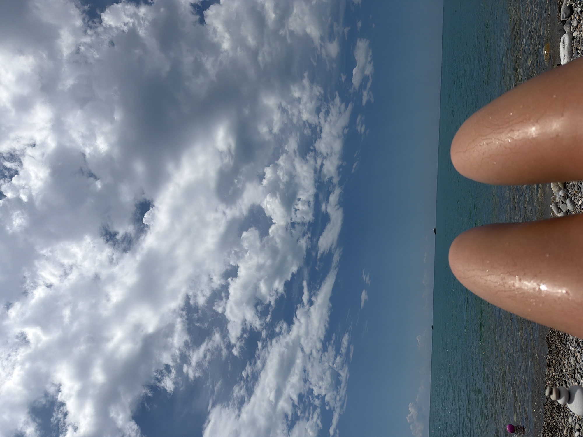 A first-person perspective shot shows a person lying on a pebble beach. The person's legs are in the foreground, with their skin glistening under the sunlight. The sea stretches out to the horizon, its surface rippling gently under a partly cloudy sky. The clouds are a mix of white and gray, casting subtle shadows on the blue sky. In the distance, small boats can be seen on the water. The beach is composed of various sizes of pebbles, and to the left, a pink ball and a stack of balancing stones are visible. The image evokes a sense of relaxation and a typical summer day by the sea. This scene is consistent with the coastal areas of Sochi, Russia, known for its pebble beaches and Black Sea views.
