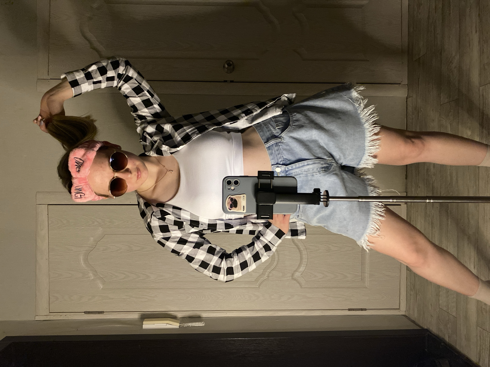 An individual is standing indoors, facing forward, with a light-colored wooden door featuring carved wavy lines and a silver hinge behind them on the left, and a darker door with a round doorknob partially visible on the right. A white wall-mounted intercom with a coiled cord is on the left wall. The floor is covered with light-toned horizontal wooden planks. The individual is wearing a white cropped tank top, an open black and white buffalo plaid button-up shirt, light blue denim shorts with frayed hems, and light-colored ankle socks. A pink plush headband with "OMG" text is worn on their head, and round, dark-lensed sunglasses with gold frames obscure their eyes. A thin gold necklace with a small crucifix pendant is visible. Their left hand is raised, holding a section of their brown hair. A grey smartphone, housed in a light-colored case featuring a cartoon pug wearing sunglasses and partially visible "SNM" text, is mounted vertically on a black clamp attached to an extendable metallic tripod, positioned in front of the individual's torso.