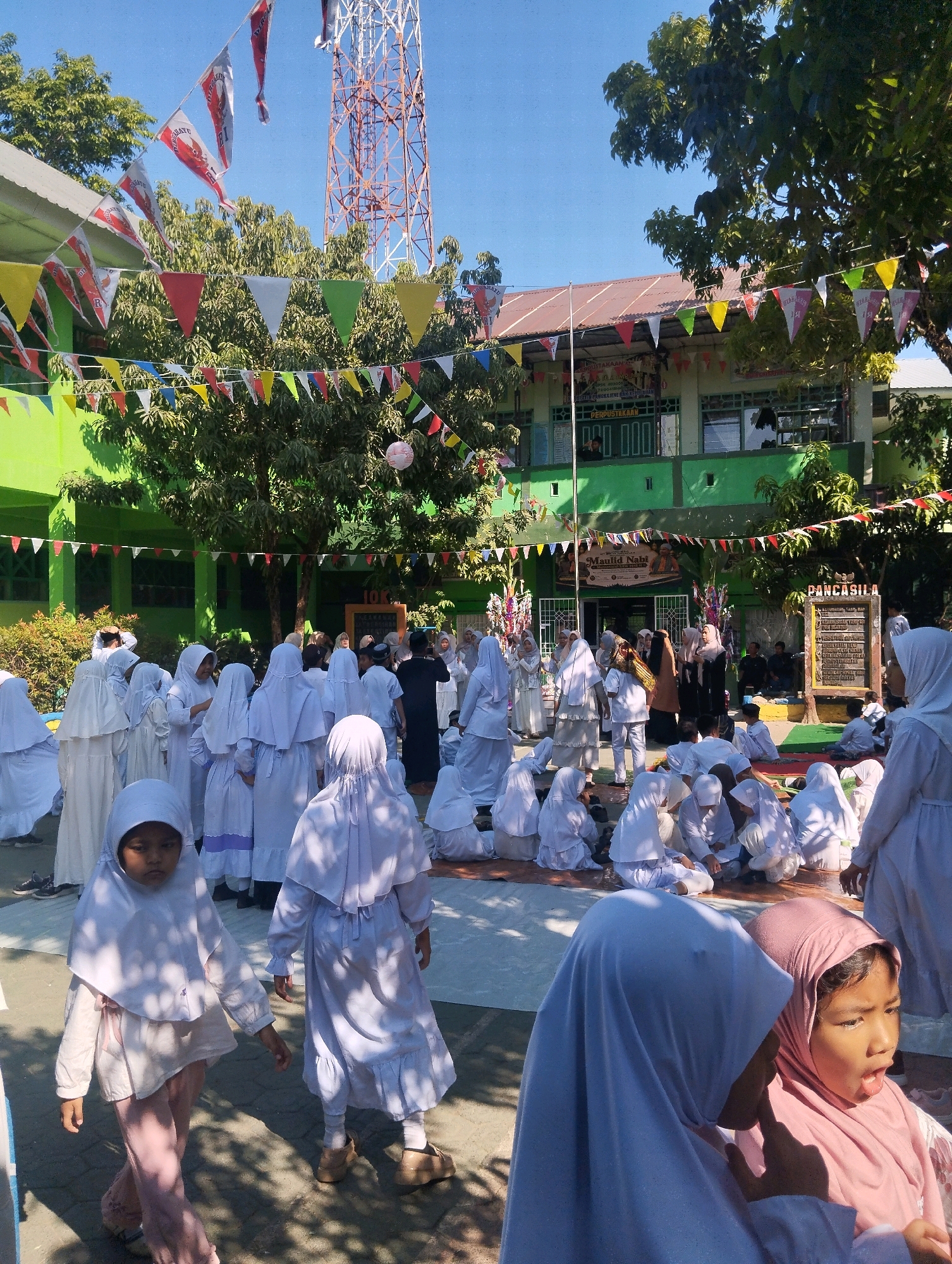 The image depicts an outdoor event, likely a school gathering, at a building in Pangkajene, Indonesia, identifiable by signage. The building, painted green with a brown roof, appears to be a school or community center. It is decorated with numerous colorful triangular flags and banners, including flags with red and white stripes, and a large banner with text that appears to be in Indonesian, possibly related to an event. A tall communication tower is visible in the background.
The foreground and middle ground are populated by a large group of children, predominantly girls, dressed in white Islamic attire, including hijabs. Some are standing, while others are seated on the ground. Several adults are also present, observing the children or participating in the event. One young girl in the immediate foreground is wearing a white hijab and a light pink outfit, walking towards the viewer. Another girl in the far foreground is wearing a pink hijab and looking to her right. The ground is paved with interlocking bricks. The atmosphere suggests a festive or celebratory occasion, possibly related to a religious or cultural event, given the attire and decorations. Sunlight casts shadows, indicating it is daytime.