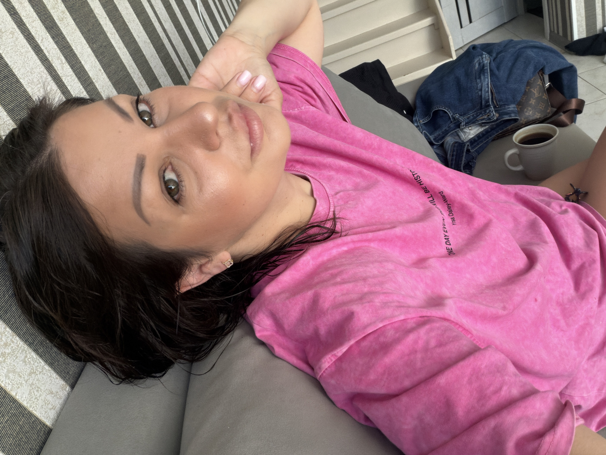A woman lies on a striped couch, her head resting on her arm. She is wearing a pink t-shirt with writing on it. A pair of jeans, a purse, and a mug of coffee are on the couch next to her. The image appears to be taken indoors, likely in a residential setting. There is no discernible connection to the city of Sochi, Russia.