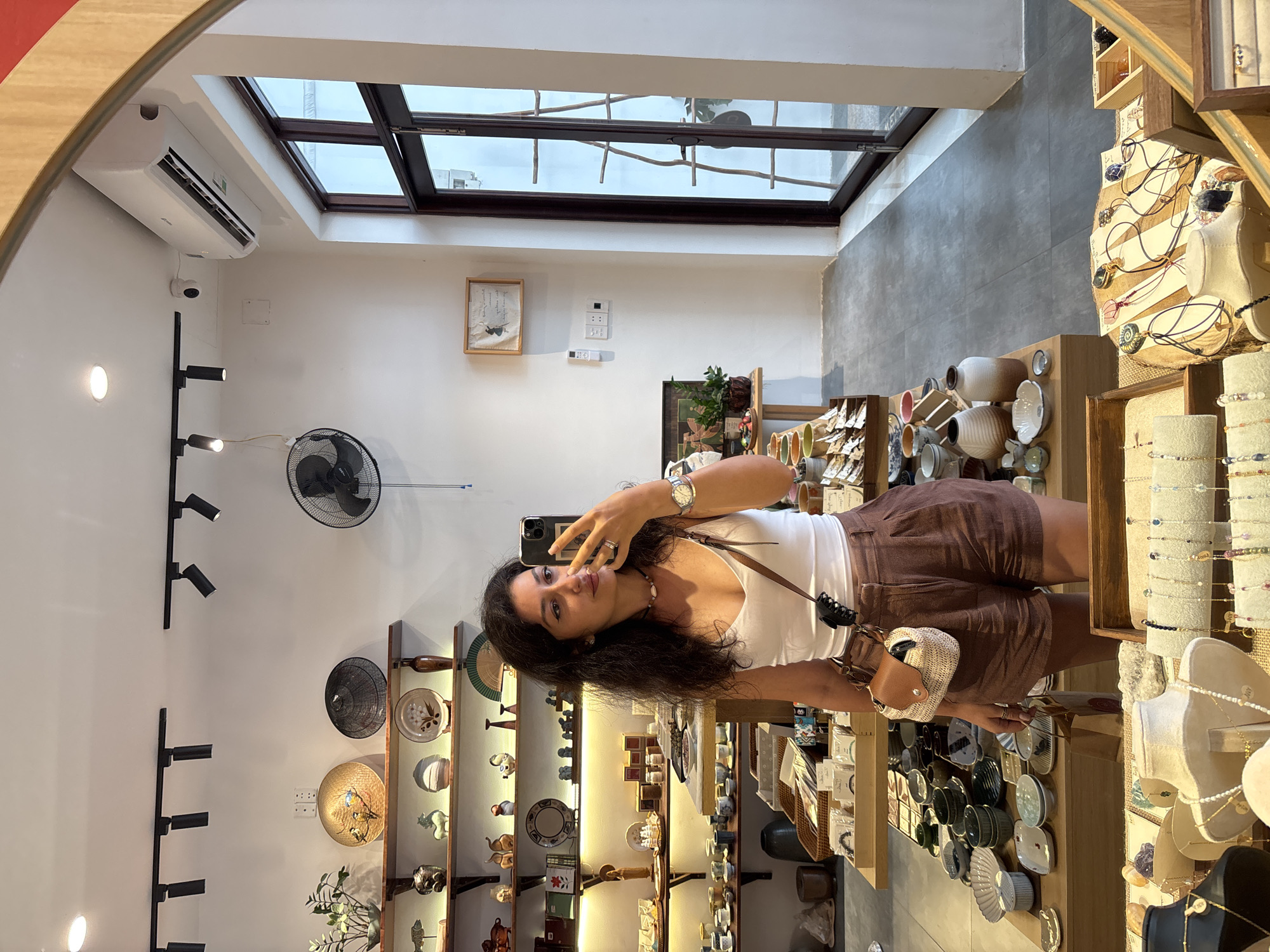 A woman stands in a well-lit shop in Đà Nẵng, Vietnam, holding a smartphone and taking a selfie. She is fair-skinned, with long, dark, curly hair and wears a white top, brown shorts, and a small purse slung over her shoulder. The shop interior has white walls, track lighting, and a skylight. Shelves display various ceramic items, woven goods, and decorative objects. A fan is mounted on the wall. The woman appears to be a customer browsing the merchandise.