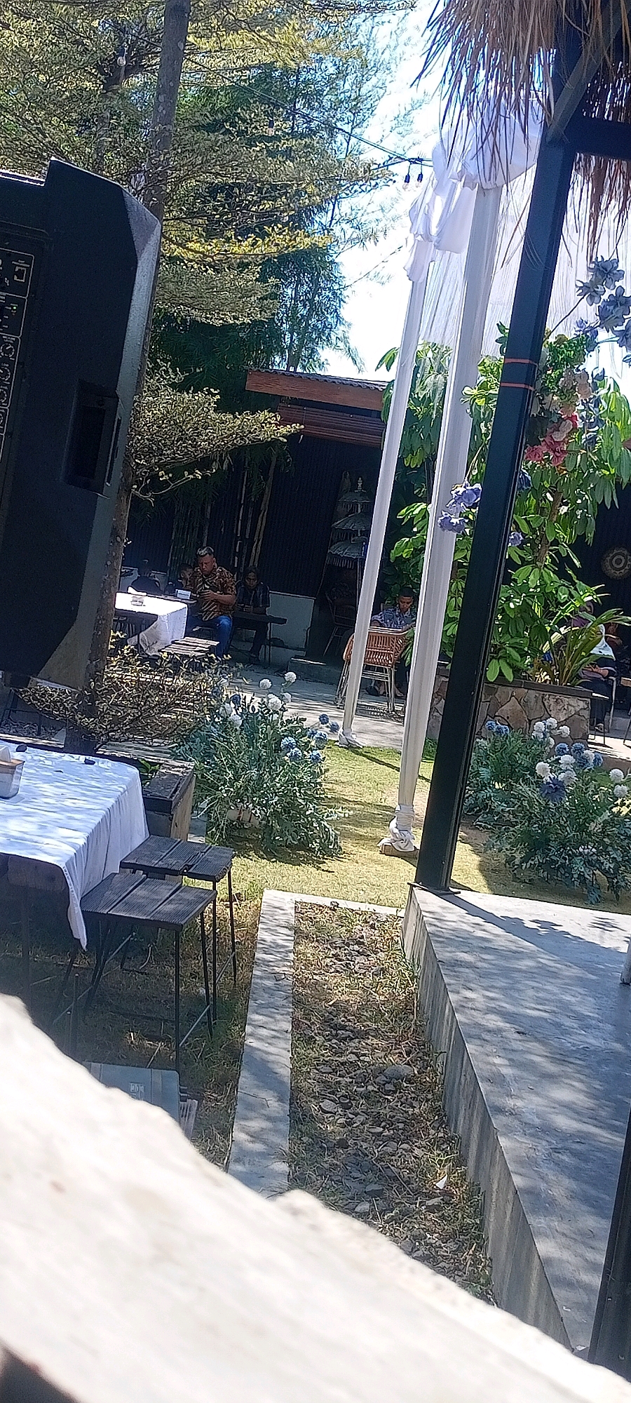 The image depicts an outdoor setting, possibly a garden or event space, in Desa Ponokawan, Indonesia. In the foreground, a large black speaker is visible on the left. To the right, a portion of a thatched roof structure with white drapes and blue flowers suggests a festive or decorative element. The middle ground reveals several tables set with white tablecloths, and wooden seating. People are seated at these tables, engaged in conversation or activities, with one individual in the center appearing to be looking at a phone. The background is characterized by lush green trees and a building with a dark facade. The ground is covered with grass and small stones, with a concrete pathway running through it. The overall scene suggests an outdoor gathering or event in a tropical or semi-tropical environment.