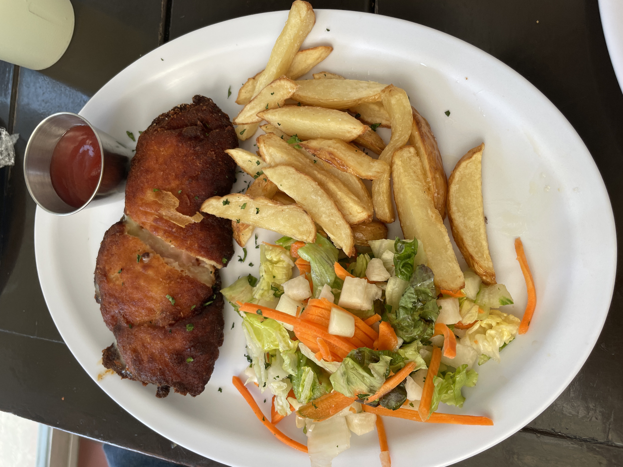 A white plate sits on a dark, possibly wooden, surface. The plate contains a meal consisting of a fried, golden-brown patty that appears to be cut open, a serving of thick-cut french fries with a sprinkle of green herbs, and a mixed salad composed of lettuce, shredded carrots, and diced white vegetables. A small, silver metal container filled with red sauce (likely ketchup) is positioned near the patty. The food arrangement suggests a casual dining setting. The location cannot be determined with certainty, but given the image content, it is unlikely to be in Mexico City, Mexico.
