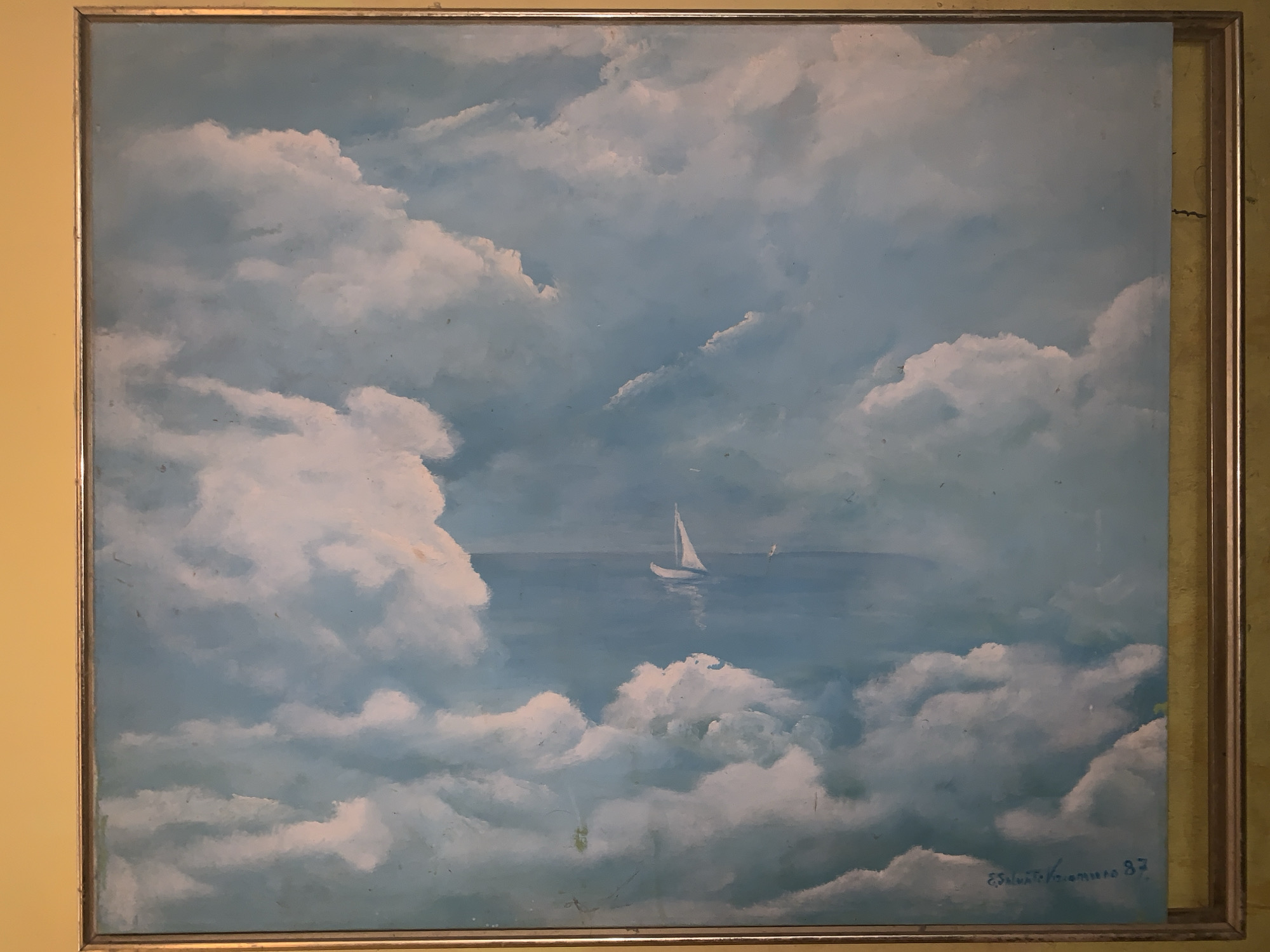 The image shows a framed painting. The painting depicts a seascape with a sailboat in the distance. The foreground is dominated by fluffy white clouds against a blue sky/sea. The frame is silver and rectangular. The bottom right corner of the painting has a signature "Eduarto V. Romero 87". The background wall is yellow. The location in the city of Santa Ana, Venezuela is not apparent from the image.