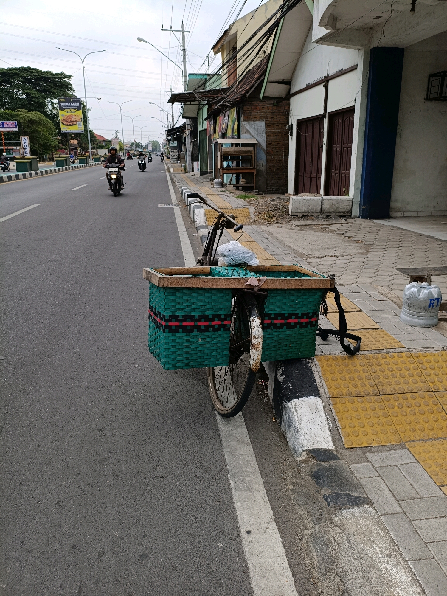 This is a medium-angle, eye-level, outdoor, daytime photograph taken on a paved road in Sewon, Indonesia. The main subject is a bicycle parked on the side of the road with a large, green woven basket attached to the front. The bicycle has a rusty wheel visible and the basket is decorated with red and black stripes. Behind the bicycle, a paved sidewalk with tactile paving and a concrete curb is visible. The road is asphalt and marked with white lines. In the background, several motorcycles are visible on the road, some with riders. Buildings line the right side of the street, featuring brown doors and architectural elements. Electrical poles and wires are present in the sky. A large banner with Indonesian text and an image of food hangs from a pole on the left side of the road. The sky is overcast.