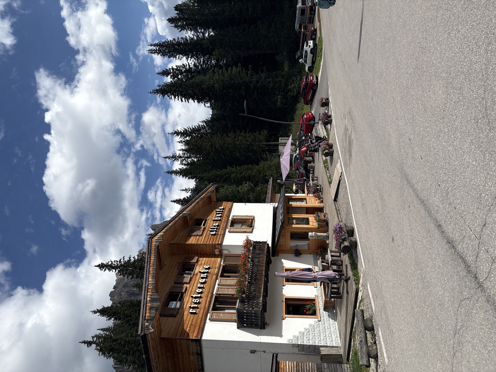 The image captures a view of the Ristorante alla Baita, situated in Auronzo di Cadore, Italy, likely at a mountain pass or a scenic location given the surrounding pine forest and mountainous terrain visible in the background under a partly cloudy sky. The building is a two-story structure with a white base and a wooden upper floor, featuring balconies adorned with red flowers. The ground floor has an outdoor seating area with wooden tables and chairs, some covered by a purple umbrella. Several cars are parked in the vicinity, including a white Jeep, a dark SUV, a red car, and a van. The paved road in the foreground leads away from the restaurant. A person is partially visible near an open doorway on the ground floor. The environment suggests a rustic, possibly tourist-oriented establishment in a natural setting.