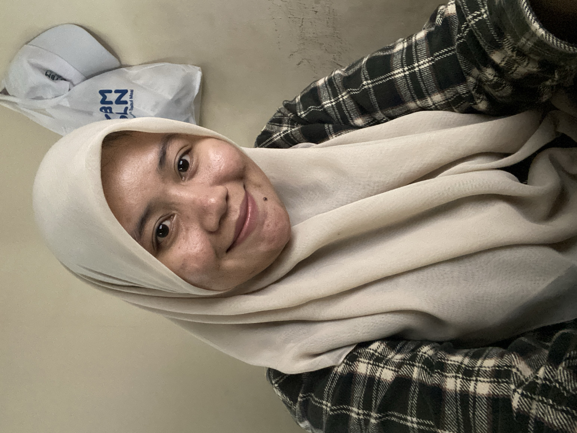 A woman wearing a light-colored hijab and a plaid shirt smiles at the camera. She has a small mole on her chin. Behind her, on a plain wall, a white plastic bag with blue lettering and a white cap are hanging. The lighting is soft and appears to be from an artificial source, illuminating the subject and creating subtle shadows on the wall. The overall impression is a casual, indoor portrait, likely taken in a residential setting in Banjarbaru, Indonesia.