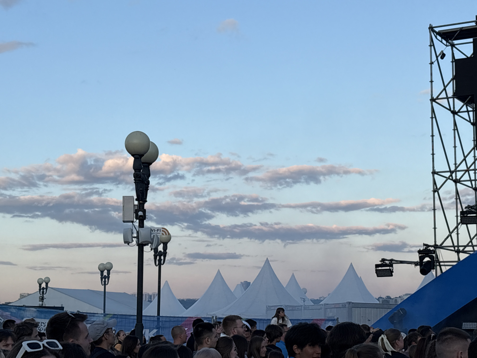 This image captures an outdoor public event, likely a festival or concert, during the late afternoon or early evening under a partly cloudy sky. The event is located in Kazan, Russia, as indicated by a partial white "K" visible on a blue banner in the mid-ground.
In the foreground, a dense crowd of people occupies the lower portion of the frame, with most individuals visible from the shoulders up. They are predominantly facing towards the right, suggesting the direction of a stage or focal point not fully visible. Individuals exhibit diverse casual attire; notable details include white-framed sunglasses on a person in the lower-left corner and a gray baseball cap on a person in the lower-center.
Dominating the mid-ground is a tall, dark pole equipped with multiple street lamp fixtures, each featuring a spherical white globe. This pole also serves as a utility mast, supporting modern communication infrastructure, including several white rectangular antenna enclosures, a dome-shaped security camera, and a sign clearly labeled "WI-FI". Additional, less prominent, similar street lamps are visible further back on the left.
The background features a series of large, white, conical-shaped tents, at least six of which are clearly visible, indicative of temporary event structures. A long, blue barrier with partially obscured white text runs horizontally behind the crowd and in front of these tents. To the far right, a substantial, dark, open-lattice metal truss structure, characteristic of stage rigging, rises vertically, supporting various lighting fixtures. The upper portion of a blue, angled surface, likely part of a stage or a large screen, is visible at the bottom right of this structure. The sky above is a blend of pale blue at the zenith, transitioning to a soft, warm, almost pink hue towards the horizon, with scattered, horizontally elongated clouds.