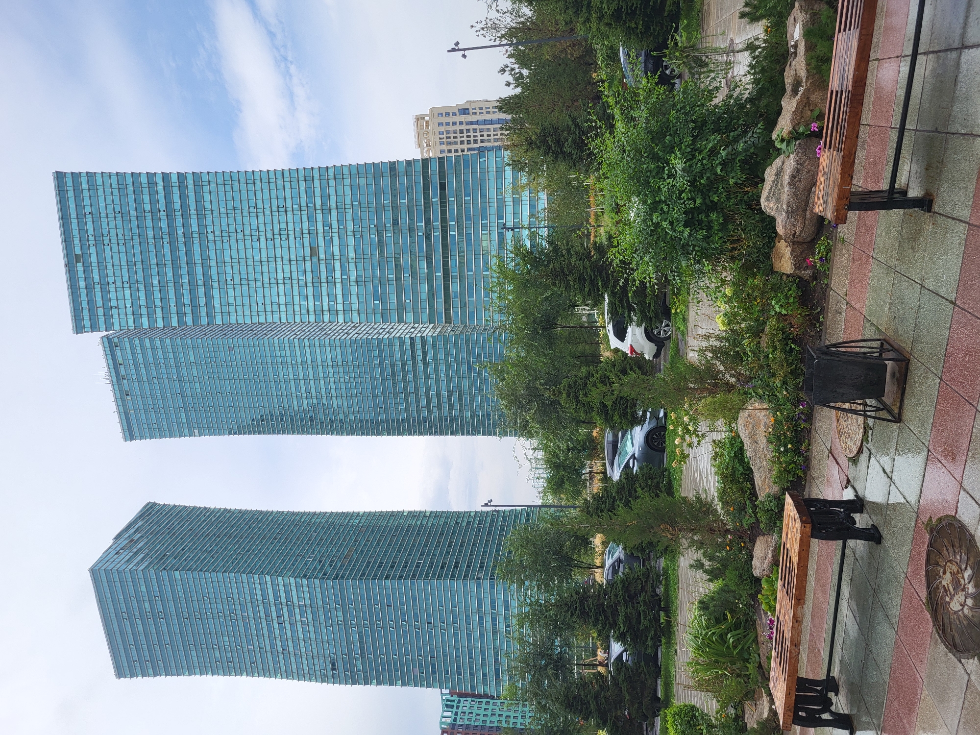 In Astana, Kazakhstan, three modern skyscrapers with a distinctive blue-tinted glass facade dominate the background. Two of these skyscrapers appear to be leaning slightly, adding a unique architectural element to the cityscape. In the foreground, a landscaped area features lush green trees and shrubs, with a paved walkway and two wooden benches. The ground is wet, suggesting recent rain. Several cars are parked along the street behind the landscaping. The sky is cloudy with patches of blue, indicating an overcast day. The scene captures a contemporary urban environment with a focus on modern architecture and public green spaces.