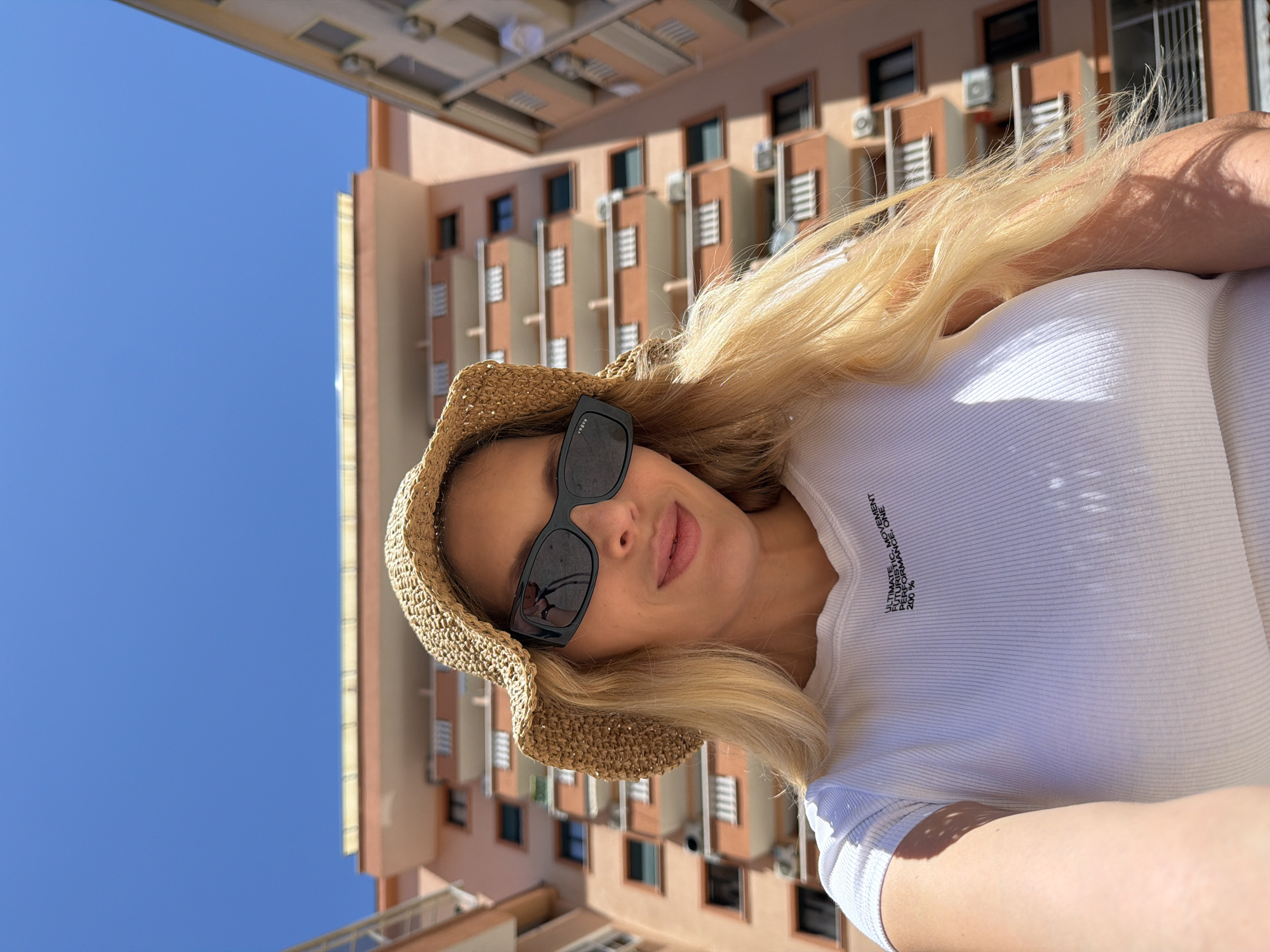 A medium shot is taken from a low angle, looking up at a blonde woman with fair skin. She is wearing a straw sun hat, dark sunglasses, and a white ribbed t-shirt with text that reads "ULTIMATE FUTURISTIC MOVEMENT PERFORMANCE. ONE 200%". The woman is looking towards the camera with a slight smile. She is outdoors, with a clear blue sky above. In the background, there is an orange apartment building with balconies and air conditioning units. The overall lighting suggests a sunny day.