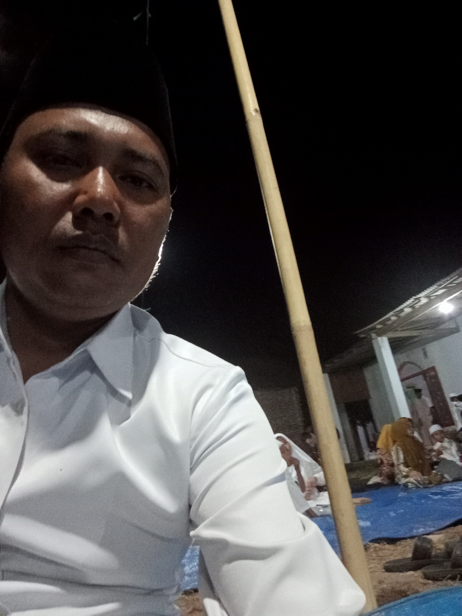 The image is a selfie taken at night in Blora, Indonesia. The foreground features a man wearing a white collared shirt and a black peci. His face is partially illuminated, showing his skin texture and some sweat. A long, light-colored bamboo pole is visible to his right, extending vertically through the frame. In the background, there are several people sitting on a blue tarp. One person wears a yellow hijab, and another a white peci. A building with a corrugated roof and lights is also visible in the background. The overall scene suggests an outdoor gathering or event at night.