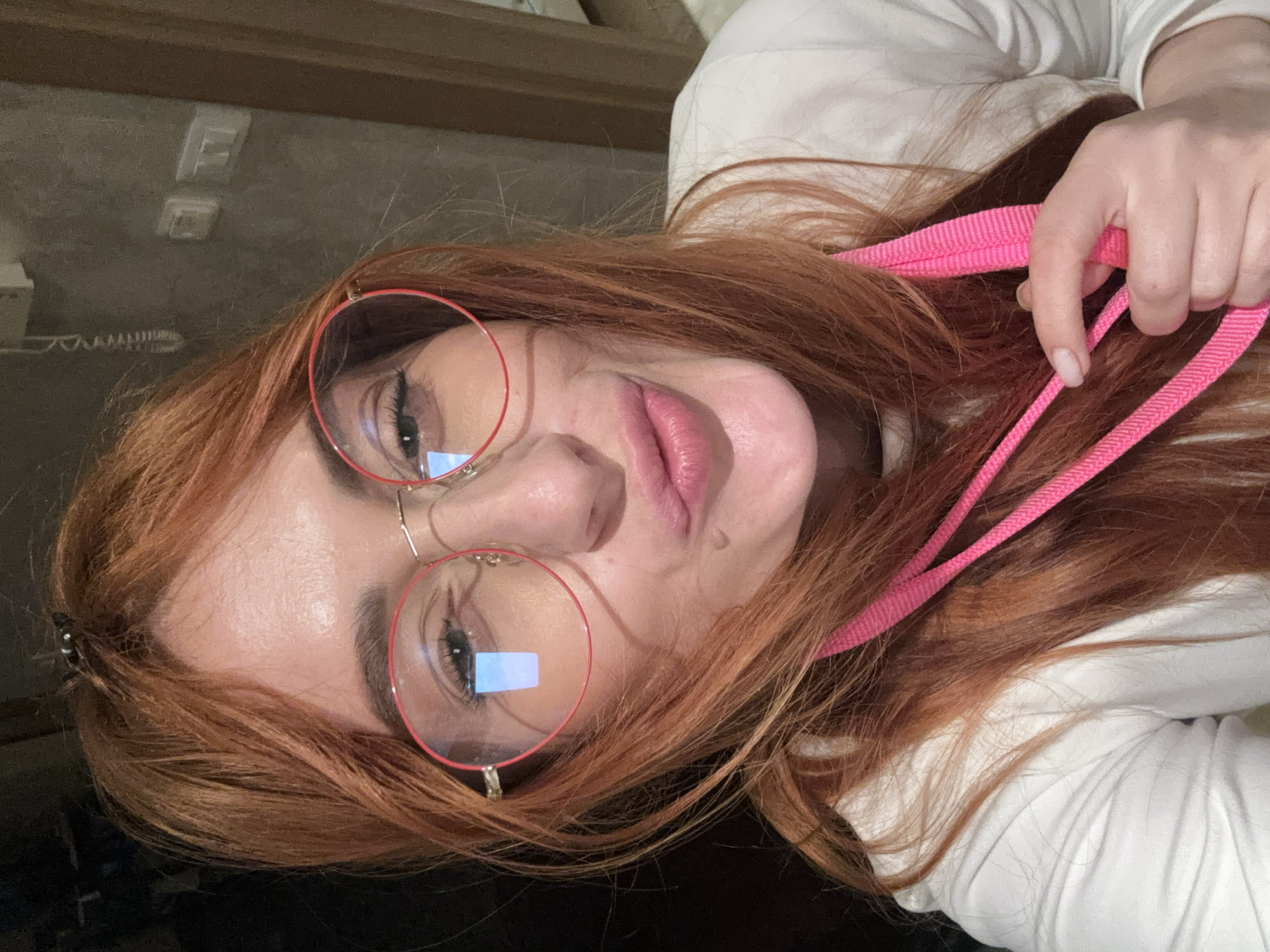 The image displays a close-up portrait of a female subject within an indoor environment located in Budva, Montenegro. The subject possesses long, auburn hair, partially secured at the crown with a small dark hair clip. She wears round eyeglasses featuring red frames and gold-toned temple arms; a distinct blue rectangular reflection, indicative of a screen, is visible in her left lens. Her facial expression presents pursed lips in a pouting gesture, and a small dark mole or blemish is present on her left cheek below the eye. She is clad in a white long-sleeved top. Her right hand, with nails painted a light, neutral color, holds a segment of a bright pink, ribbed strap or lanyard. The background reveals a dark gray, textured wall behind the subject's left side, featuring a white intercom-like device with a coiled cord positioned above and to the left of a white double electrical switch plate. A vertical light-colored wooden architectural element, possibly a door frame, is partially visible on the right side of the image. The scene is illuminated by artificial light sources.