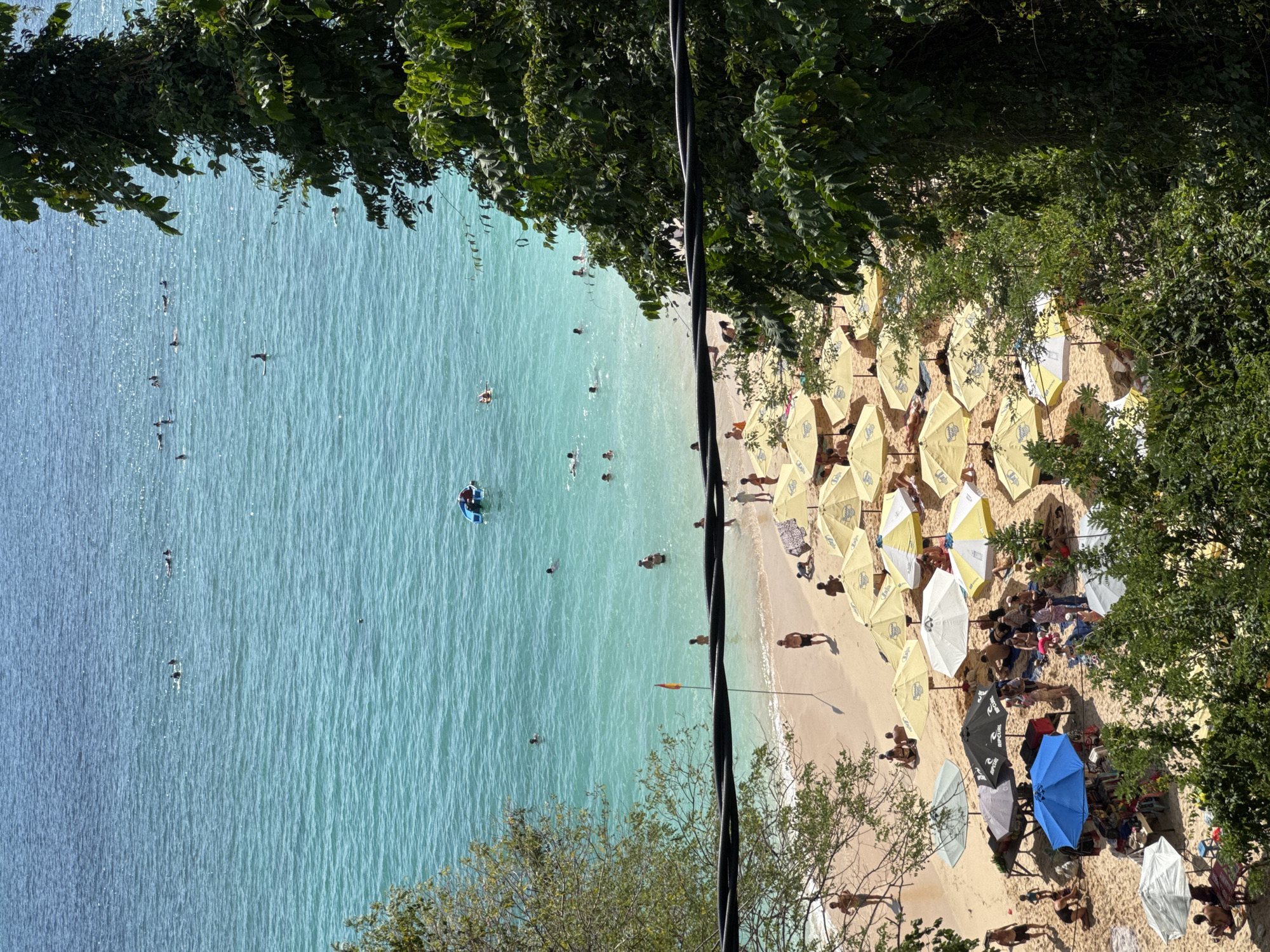 The image depicts a crowded beach scene in Kuta Selatan, Indonesia, on a sunny day. The turquoise water is populated with numerous people swimming, surfing, and enjoying various water activities. Some are riding surfboards, while others are simply floating or swimming. A small blue inflatable raft is visible in the water.
The sandy beach is covered with many yellow and white umbrellas providing shade for beachgoers. People are sunbathing, sitting under the umbrellas, and walking along the shore. Several beach vendors are present, with stalls set up selling goods, indicated by the presence of umbrellas and tables laden with items. A red and yellow flag is planted on the beach, possibly indicating a swimming area or warning.
Lush green trees and foliage frame the scene from the sides, with a prominent dark cable running horizontally across the foreground, partially obscuring the view. The overall impression is a vibrant and active beach environment characteristic of a popular tourist destination.