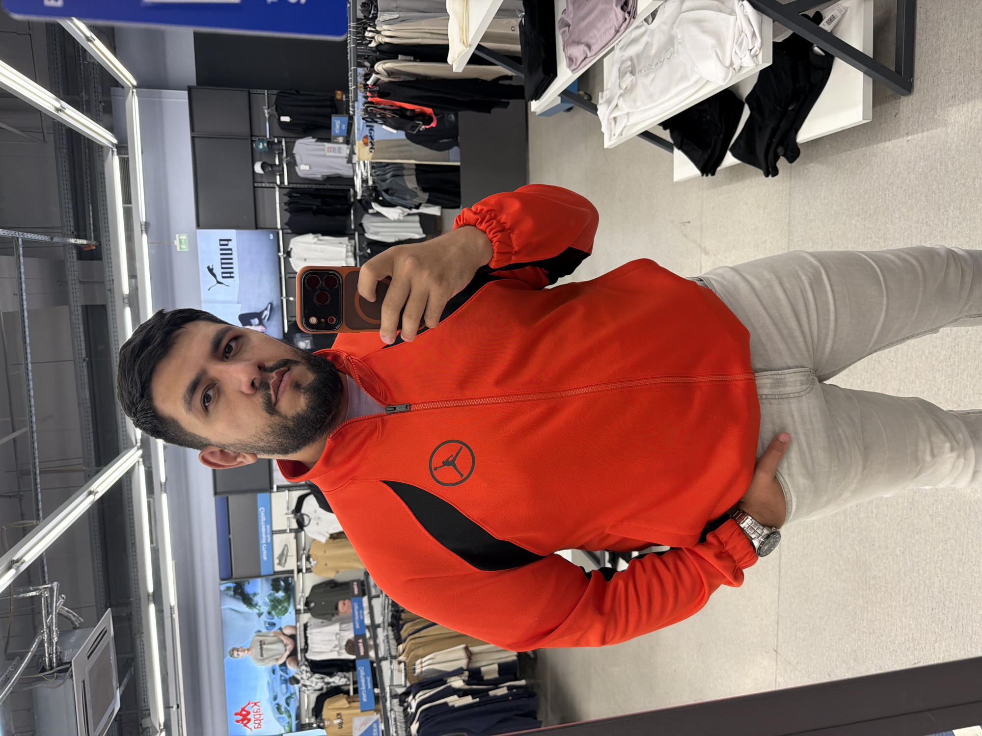 A man is taking a selfie in what appears to be the mirror of a dressing room or fitting room inside a clothing store. He is wearing an orange Jordan brand jacket with black accents, light grey pants, and a silver wristwatch on his left wrist. The man is holding a brown iPhone to take the picture. He has dark hair and a beard. Behind him, clothing racks are visible, displaying a variety of garments. The store appears to be well-lit with fluorescent lighting. Signage for Puma and Kappa can also be observed. The location is likely a sportswear store, potentially in Krasnoyarsk, Russia.