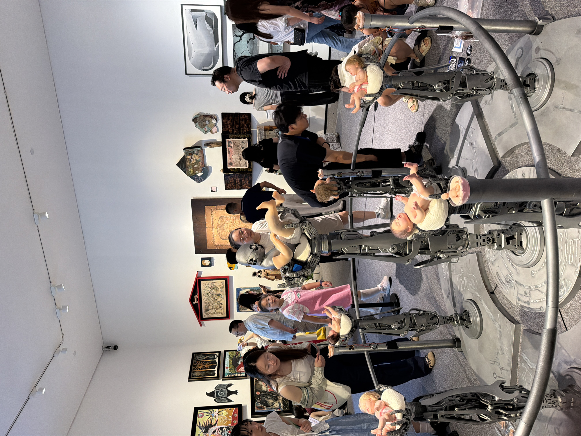 The image captures an indoor art exhibition within the Chongqing High-tech Zone, China. The scene is populated by numerous visitors observing various artworks. In the foreground, a circular art installation comprises several metallic robotic or mechanical arms, each supporting a lifelike baby doll in different infant poses. A gray metal railing surrounds this central display, separating the viewers from the artwork. Many attendees, including adults and children, are engaged with this installation; some are leaning over the railing for closer inspection, while others photograph it with their mobile phones. A young girl in a pink dress is prominently positioned near one of the baby dolls, observing it intently. The white walls of the exhibition space are adorned with a diverse collection of framed artworks, including paintings, prints, and photographs of varying sizes and styles. The ceiling features a system of track lighting with multiple spotlights illuminating the gallery. The floor is covered with gray carpeting. A security camera is visible mounted on the wall near the ceiling in the background.