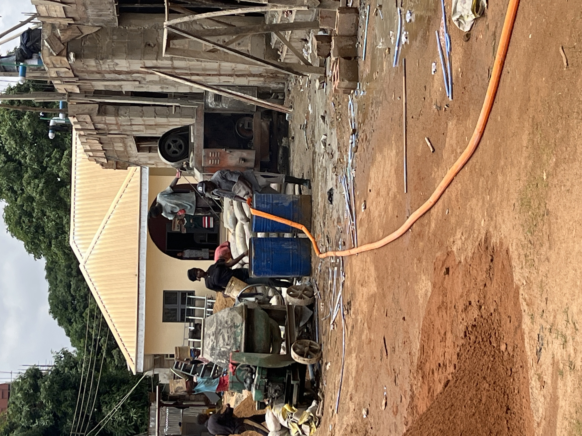 The image depicts an active construction site in Jos, Nigeria. The scene is characterized by a partially built concrete structure with scaffolding. In the foreground, several construction workers are engaged in tasks involving a cement mixer and large blue barrels. One worker, wearing a gray shirt and cap, is standing next to a barrel, holding a white bucket. Another worker in a black shirt is also near a barrel, holding a bucket. An orange hose snakes across the muddy ground, connecting to one of the barrels. There is a pile of reddish-brown dirt to the left, and various construction materials, including bags of cement or sand, are visible. In the background, a yellow-roofed building with an arched entrance stands adjacent to the construction. Lush green trees are visible behind the structures, and the sky is overcast. The ground is wet and muddy, suggesting recent rain.