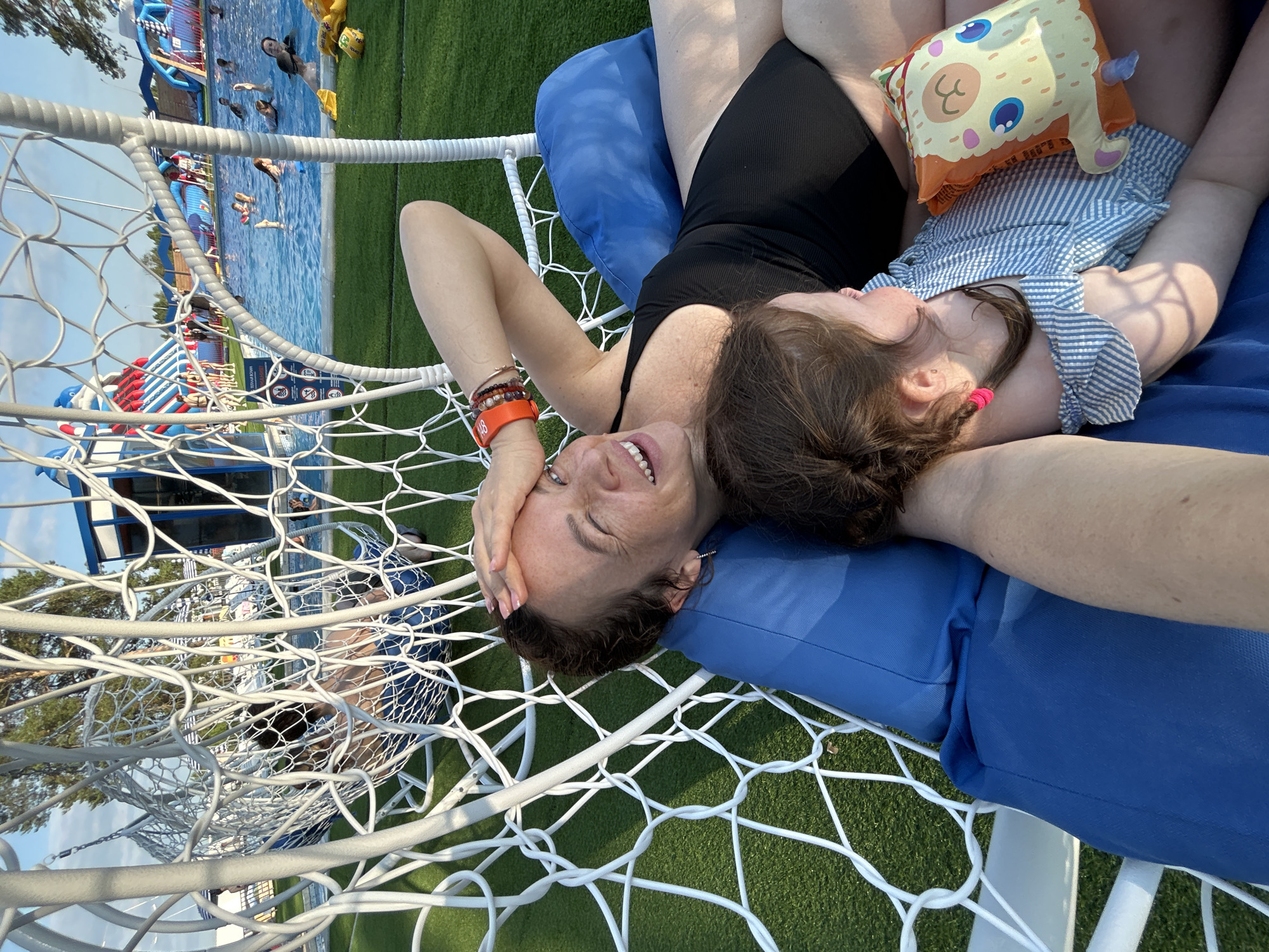 This media file features a selfie-style shot of two people, an adult and a child, lying on a blue cushioned hammock. The adult, with their hand on their forehead, appears to be smiling with their eyes closed. The child, nestled close to the adult, is wearing a blue and white striped dress and has pink hair ties. A small, cartoonish plush toy rests on the child's chest. The hammock is suspended within a white netting structure.
In the background, a swimming pool is visible with several people swimming and playing. There are inflatable water slides and structures suggesting a water park or resort. The surrounding area is covered in green artificial turf. The lighting suggests it is daytime, likely late afternoon or early evening, with the sun casting shadows. The overall atmosphere is one of relaxation and enjoyment.
Visible text includes branding on the inflatable structures and informational signs near the pool, but these are not legible enough to decipher specific details. The location context provided is Votkinsk, Russia, which aligns with the recreational outdoor setting depicted.