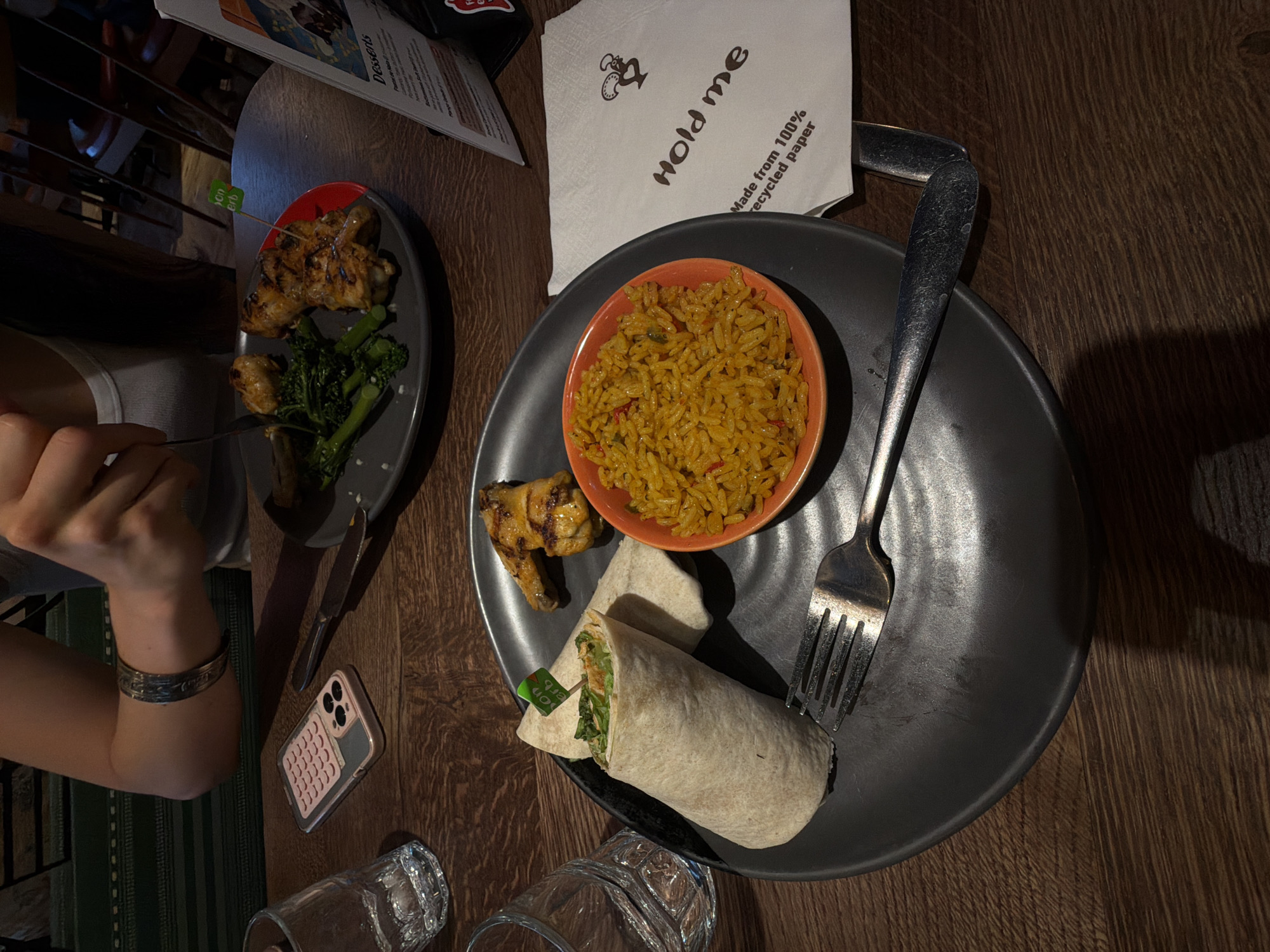 The image shows a table setting in a restaurant. The table has a dark wooden surface. There are two plates visible. The first plate, on the left, has chicken pieces and broccoli. The second plate is larger and contains a bowl of rice, a wrap, and more chicken pieces. A fork rests on this plate. A napkin with the words "Hold me, Made from 100% recycled paper" is also visible. There are also some glasses, a phone, and a menu visible on the table. A person's hand with a silver bracelet is visible. I am unable to identify the precise location in London, United Kingdom.