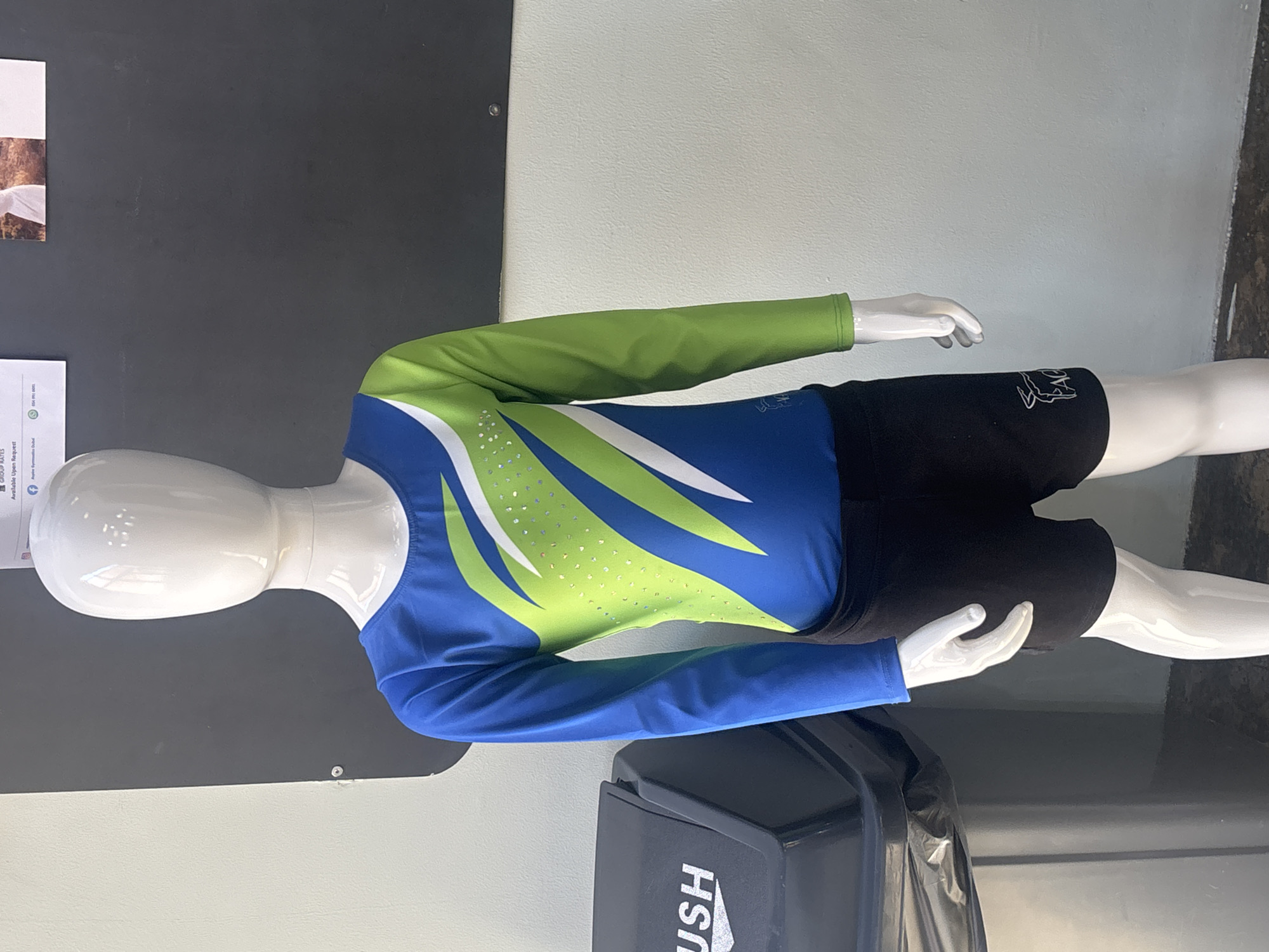 A full-body, headless white mannequin is positioned in front of a dark grey wall and a white wall. The mannequin is wearing a long-sleeved leotard with a blue top and sleeves, and a lime green and white abstract design on the front. The leotard has scattered clear rhinestones on the green design. The mannequin is also wearing black shorts with a small white logo on the left leg. To the left of the mannequin is a black trash can with the word "USH" visible on its side. In the background, a portion of a white bulletin board with some text and an image is visible on the dark grey wall. The lighting suggests an indoor environment.