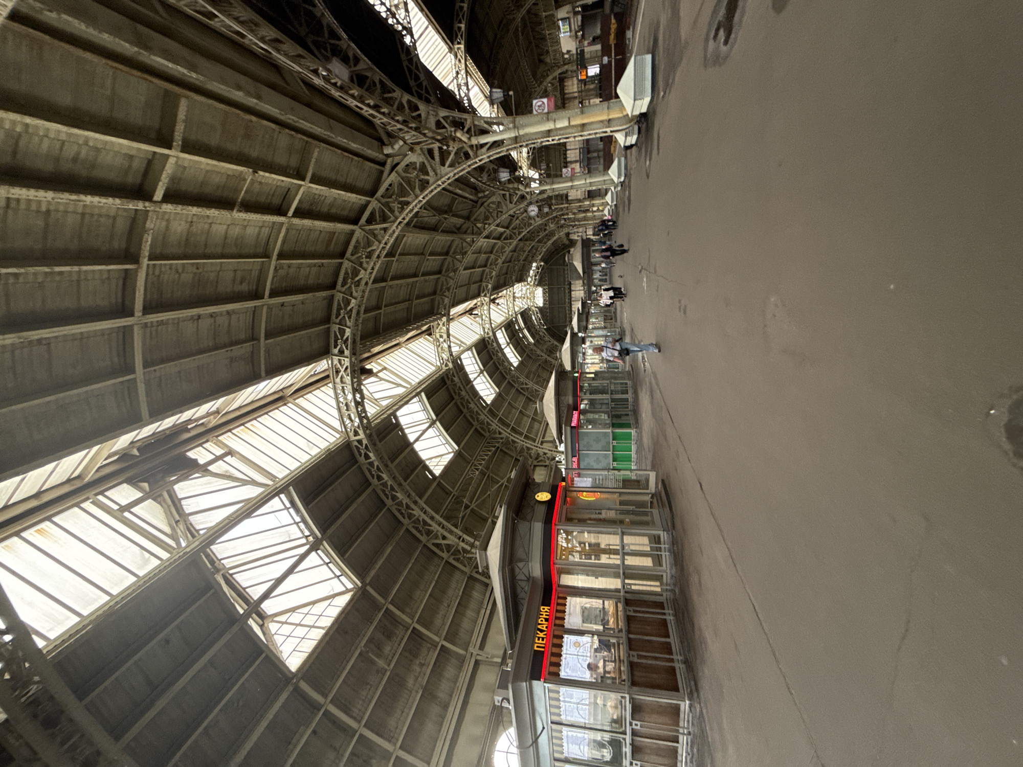 The image shows the interior of a large, historic train station in Saint Petersburg, Russia, specifically Vitebsky Railway Station. The station features a vast, arched glass and metal roof, with sunlight filtering through the translucent panels, illuminating the spacious concourse below. Several small kiosks are visible along the walkway, one of which is a bakery displaying goods and a menu. People are seen walking along the concourse, some appearing to be passengers or staff. The architecture is characterized by intricate metalwork and a grand, industrial aesthetic. The ground is paved with a neutral-colored material, and there are visible architectural supports and lighting fixtures throughout the space.