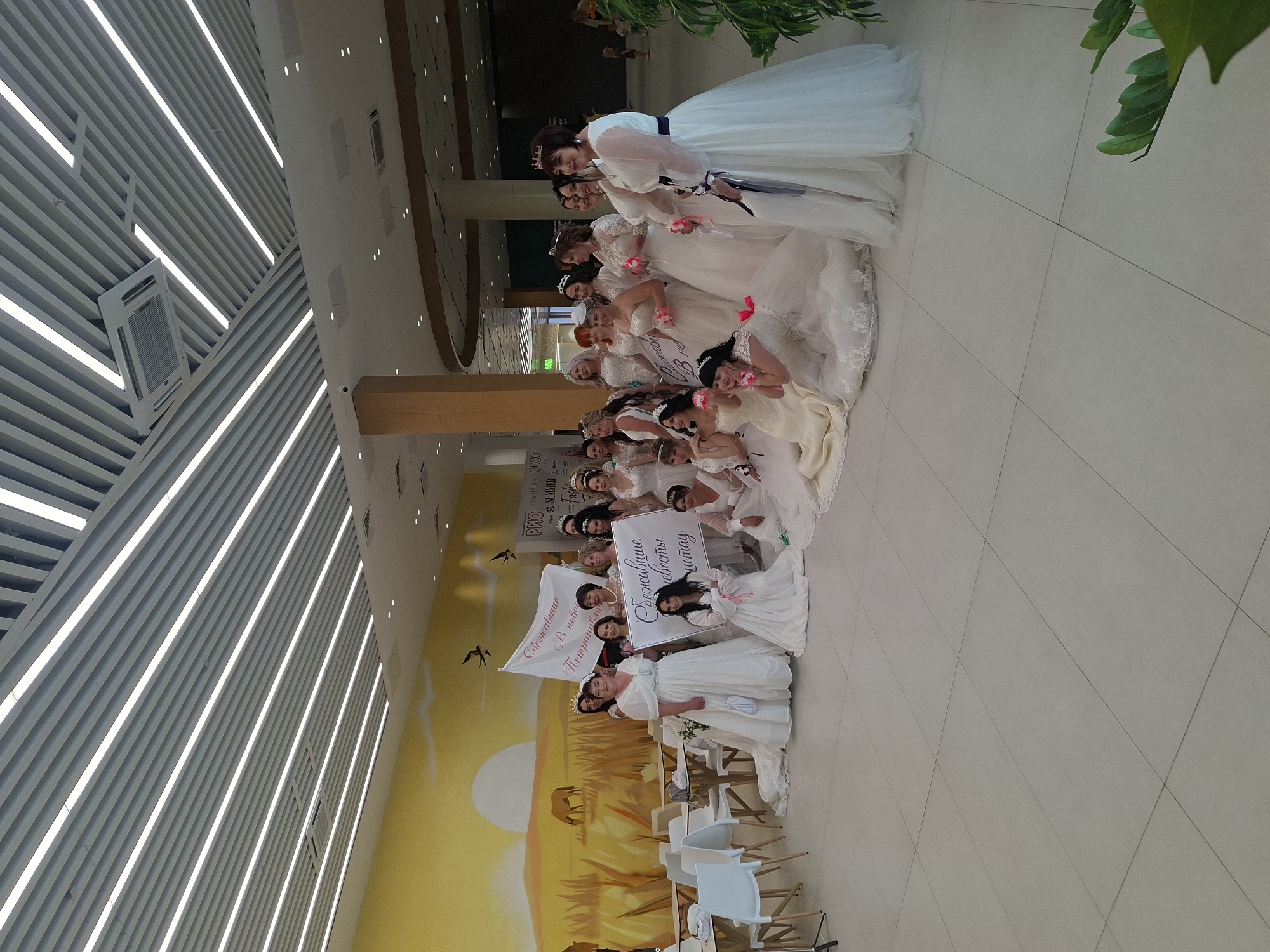 A group of women, all wearing white wedding dresses and some with crowns, are posing for a photograph in a brightly lit indoor space. The women are of various ages and ethnicities. Some are standing, while others are kneeling. They are holding two signs, one in Russian which reads "Свадебное путешествие в Петропавловск" which translates to "Honeymoon in Petropavlovsk", and another with similar text. The background includes a mural depicting a sunset over a grassy field with a horse, as well as a modern ceiling with linear lighting. There are tables and chairs visible in the foreground, suggesting a public area like a mall or event space. The floor is tiled. The lighting is even and bright, with no harsh shadows. The overall atmosphere appears festive and celebratory. The image's metadata indicates it was taken in Kokshetau, Kazakhstan.