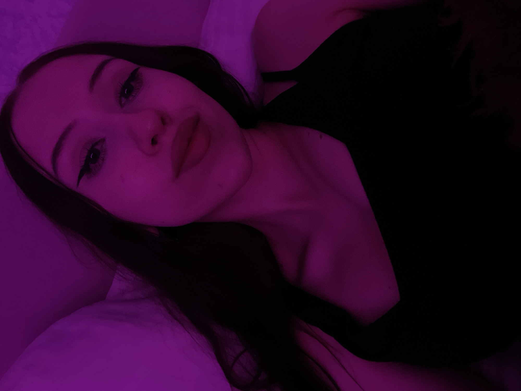 The image shows a close-up, overhead shot of a young woman lying down. She has dark hair and is wearing a black tank top. The lighting is predominantly purple, suggesting a low-light environment, possibly illuminated by a colored light source. Her eyes are open and she is looking towards the camera. The background appears to be a soft surface, likely pillows or bedding, which are also illuminated by the purple light. There is no identifiable information in the image that would suggest a location in Votkinsk, Russia.