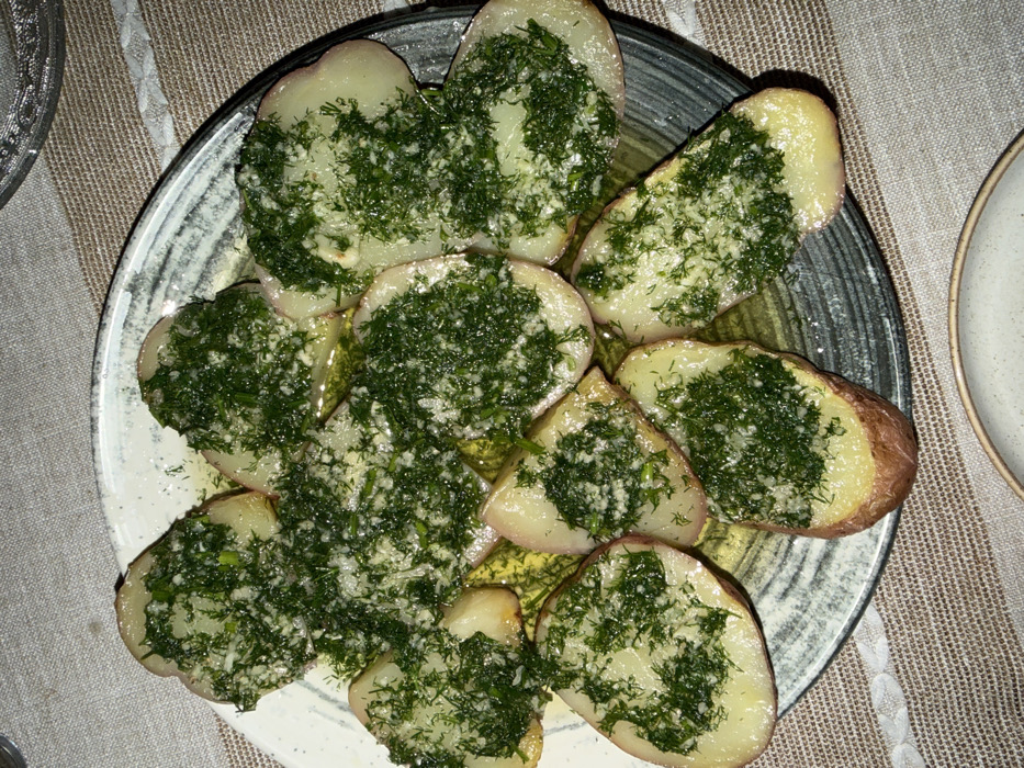 The image displays a close-up, overhead view of a plate filled with food, likely a side dish or appetizer, resting on a textured, striped placemat or tablecloth.
The main subject is a collection of potato halves, primarily showcasing their cut sides, which are light yellow in color. Many of the potatoes still have their skin on, which appears reddish-brown, suggesting they might be red potatoes or a similar variety. Each potato half is generously topped with a vibrant green mixture of finely chopped herbs, most prominently resembling dill, along with small white specks that appear to be minced garlic. The herbs and garlic are glossy, suggesting they are coated in oil, and a small pool of a clear, golden liquid (likely olive oil or butter sauce) is visible at the bottom of the plate, mingling with the potatoes.
The potatoes are arranged somewhat haphazardly on a dark-colored, round plate with a subtle speckled or mottled pattern on its surface and rim. The plate itself rests on a light beige or off-white fabric surface, possibly a placemat or tablecloth, which has faint, lighter-colored stripes running vertically.
In the upper left corner of the image, a sliver of another clear, possibly glass, plate or dish with an intricate, etched pattern is visible. On the right side of the image, a portion of a plain, light gray or off-white ceramic dish (possibly a bowl or plate) can be seen. The overall lighting appears to be indoor and soft, highlighting the textures of the food and the setting. There is no visible text in the image.