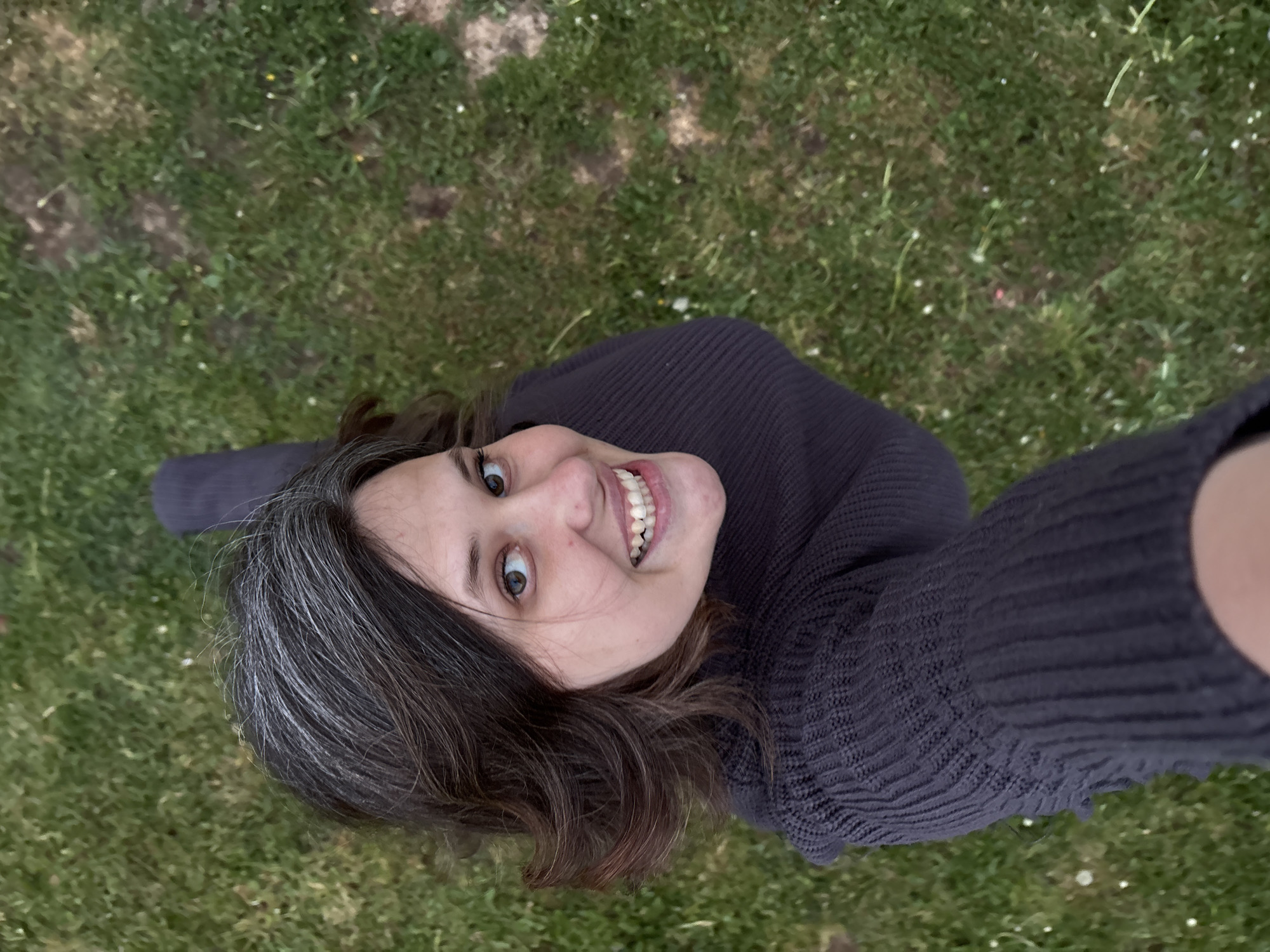 A woman is taking a selfie from a low angle on a grassy lawn. The background is filled with green grass, and there are some bare patches of dirt visible. The woman has dark brown hair with graying roots, fair skin, and is wearing a dark-colored knit sweater. Her eyes are wide, and she is smiling broadly, showing her teeth. Her head is tilted slightly to the right. It is impossible to confirm the location of the photo in Прикамский, Russia, based on the provided image content alone.
