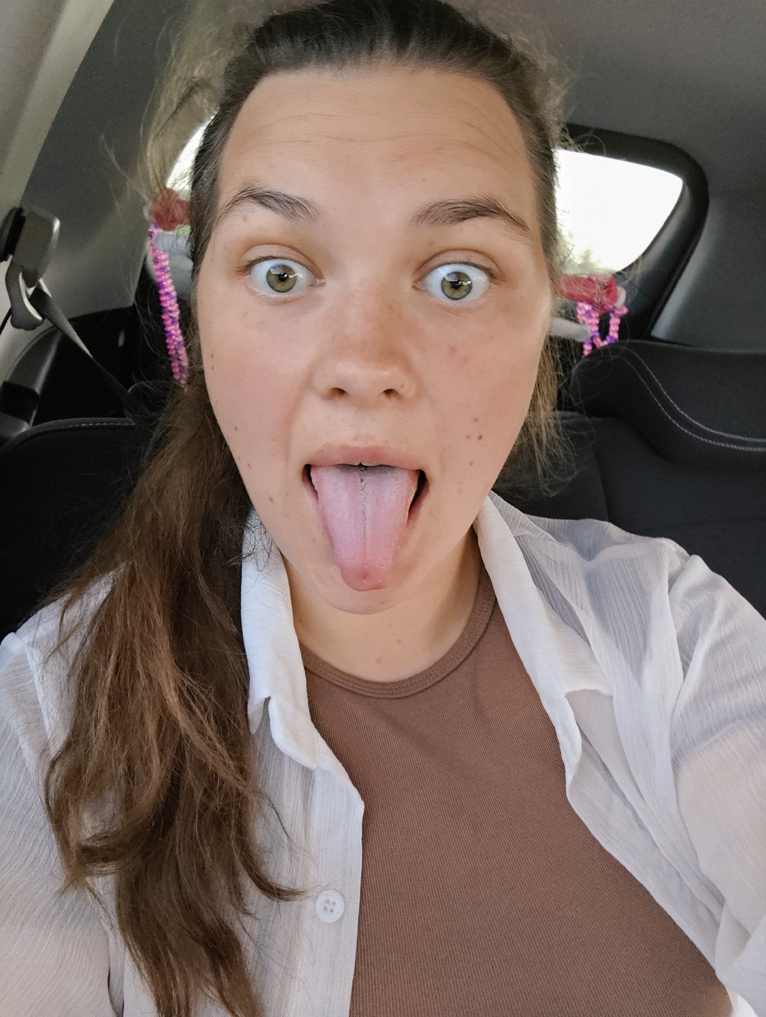 A woman is inside a vehicle. She has long, brown hair, pulled back into a ponytail with colorful decorations. She is wearing a brown shirt and a white button-down shirt. Her eyes are wide, and she is sticking out her tongue. The background suggests a car interior. The location Фоки, Russia is not discernible from the image.