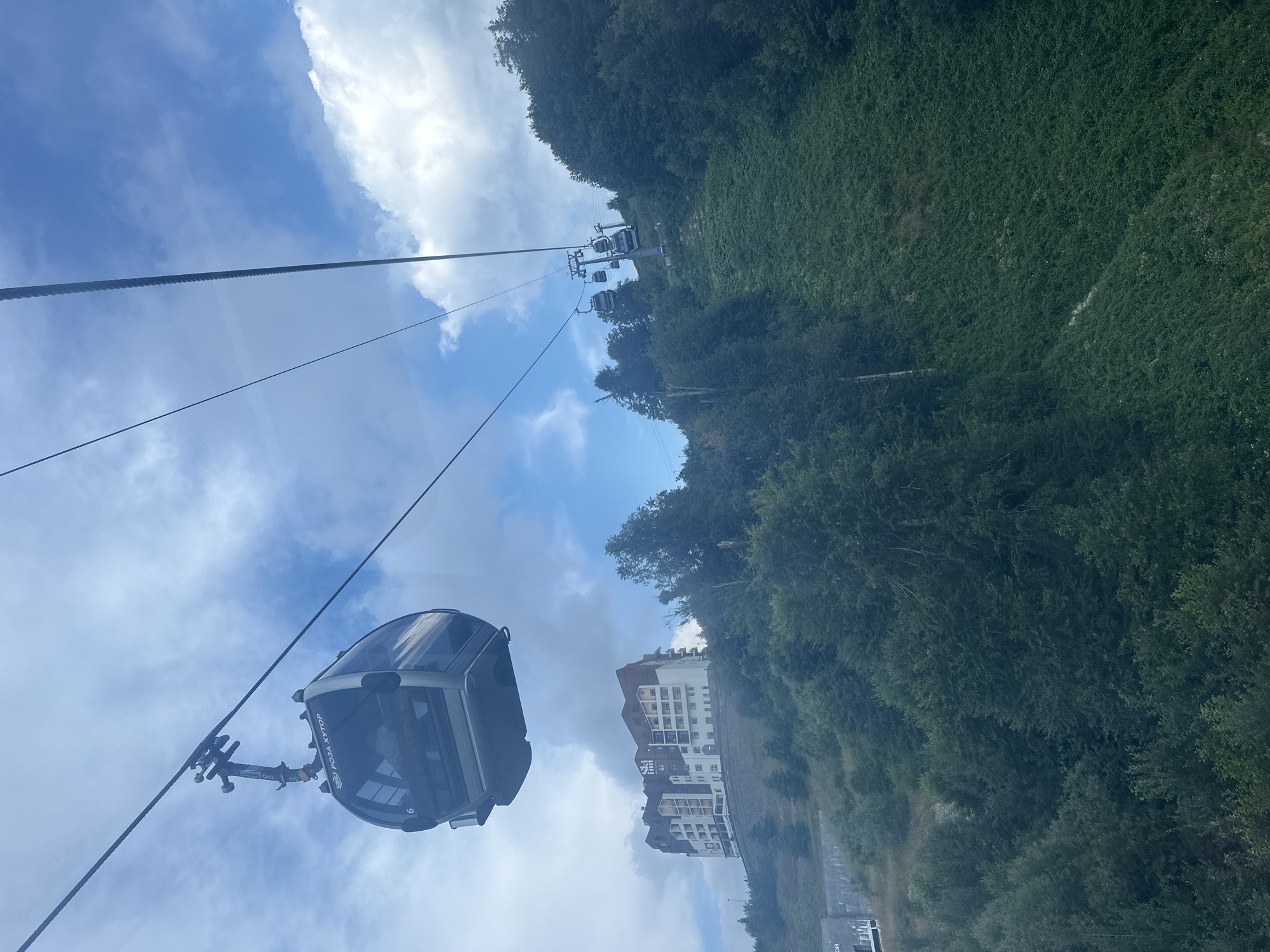 The image shows a cable car in motion, likely on a sunny day with scattered clouds. In the foreground, a modern cable car cabin, labeled "ROZA KHUTOR," is suspended from a thick cable. The cabin is dark gray with large windows and appears to be empty.
In the background, a multi-story hotel building with a modern architectural design stands on a tree-covered hill. The hotel is predominantly white with beige and dark brown accents. The hill is covered with dense green vegetation, including trees and grass. Further along the cable line, several other cable car cabins are visible ascending the slope, indicating a functioning ski resort or mountain transportation system. The environment appears to be a mountainous region with lush greenery. The location is identified as Estosadok, Russia, a popular resort area.