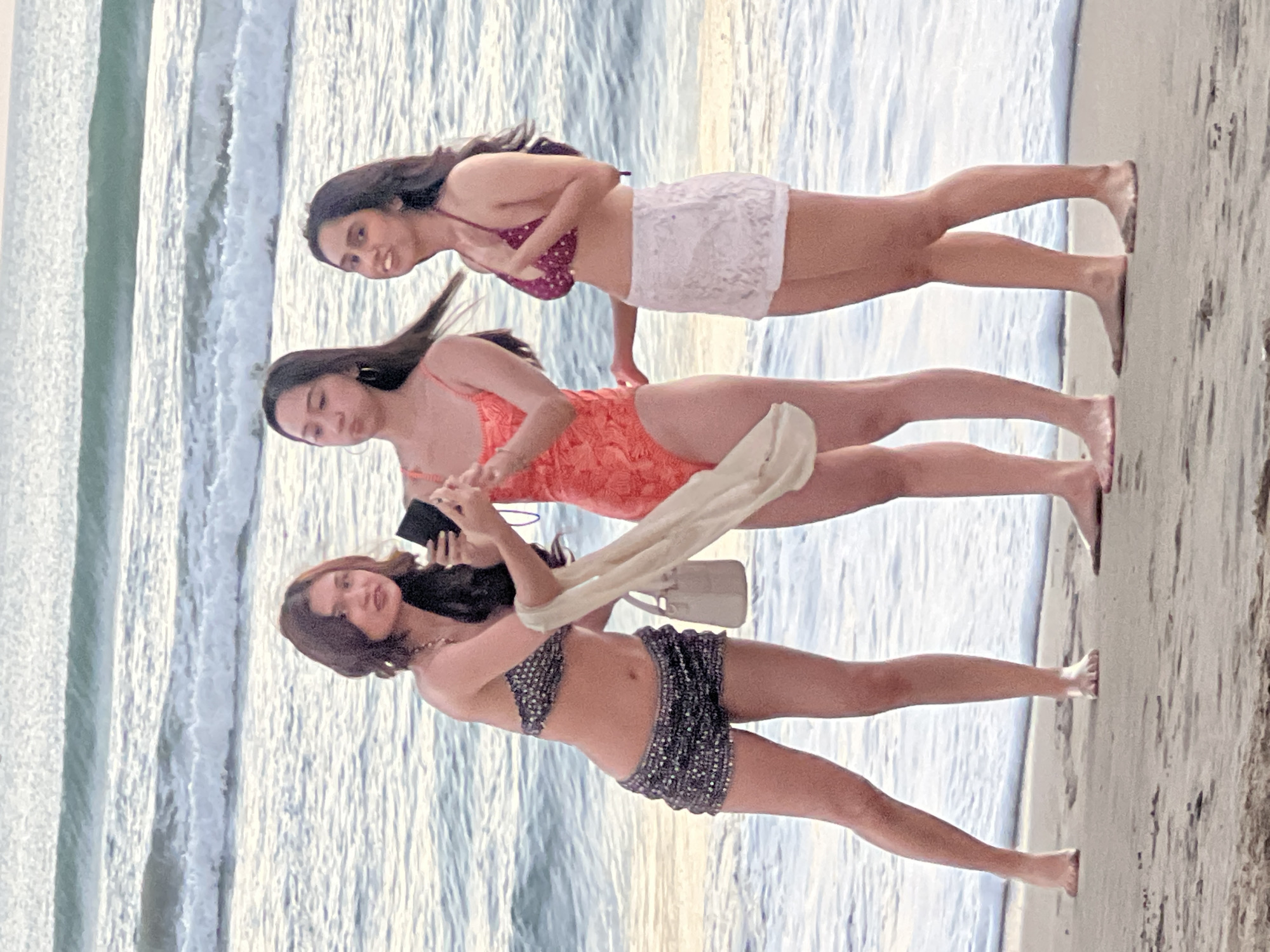 Three young women are standing on a sandy beach in Kuta, Indonesia, near the ocean. The woman on the left is wearing a sparkly bikini top and shorts, holding a tan purse and a light-colored scarf, and appears to be taking a selfie with her phone. The middle woman is wearing an orange patterned one-piece swimsuit and is looking at a phone. The woman on the right is wearing a burgundy bikini top and white lace shorts, and is smiling while looking forward. The ocean waves are visible behind them.