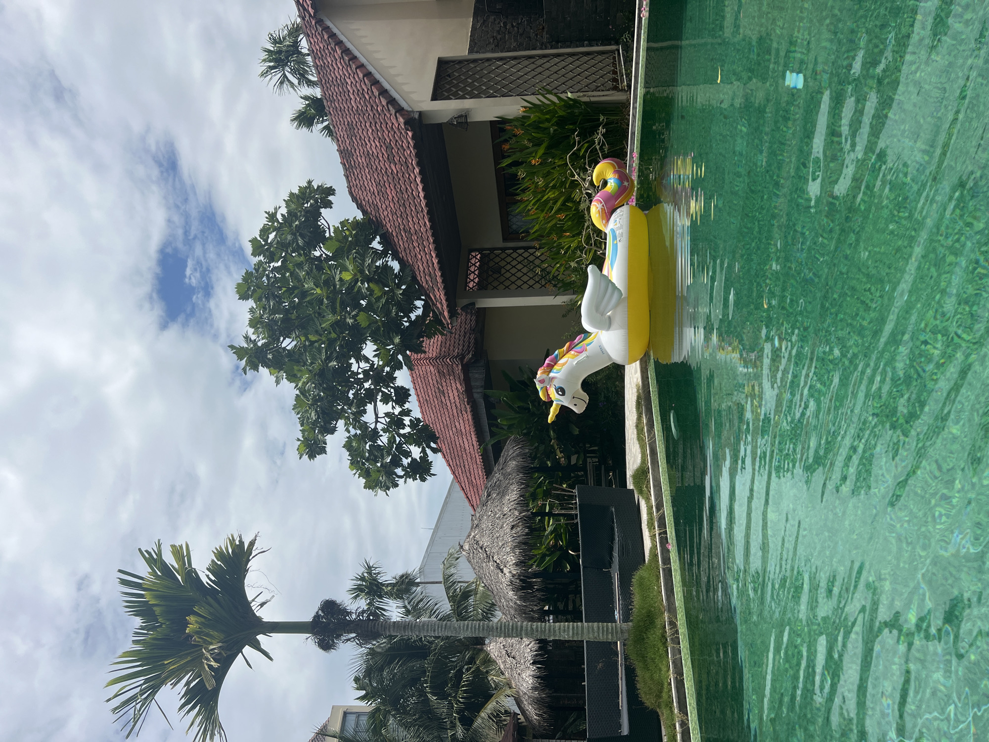 An inflatable yellow unicorn floats in a green swimming pool. The unicorn has a rainbow mane and tail, and white wings. In the background, there are two thatched-roof huts, lush green trees, and buildings with terracotta roofs. The sky is cloudy. This scene appears to be from a resort or hotel in Hoi An, Vietnam, given the architectural style and tropical vegetation.