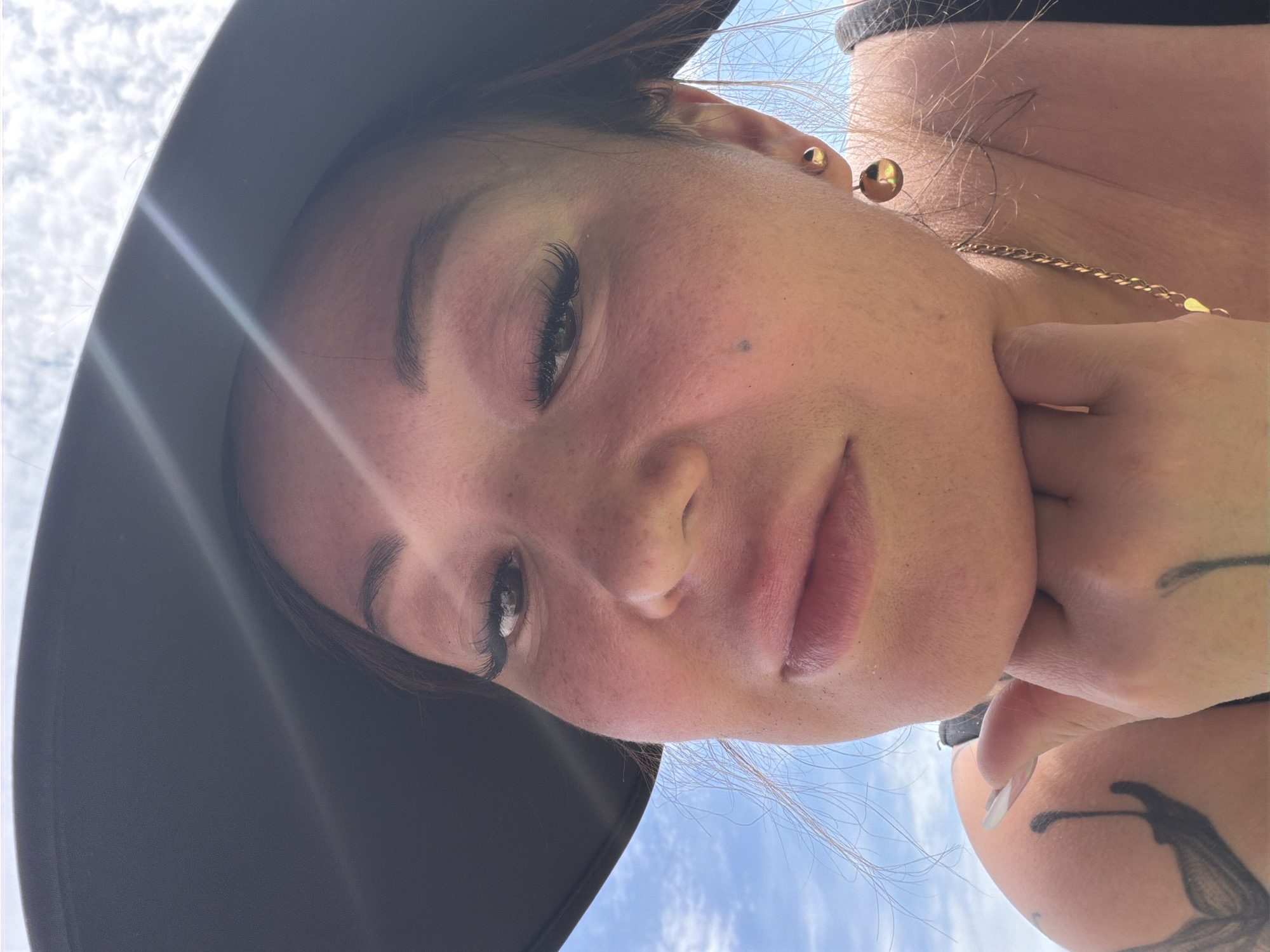 A close-up, upward-angled photograph depicts a woman with fair skin, freckles, and extended eyelashes. She is wearing a wide-brimmed hat, which casts a shadow over the upper portion of her face. She is wearing gold earrings and a gold necklace. Her right hand is visible near her face, showing a tattoo on her arm. The background features a slightly blurred blue sky. The location is not specified.